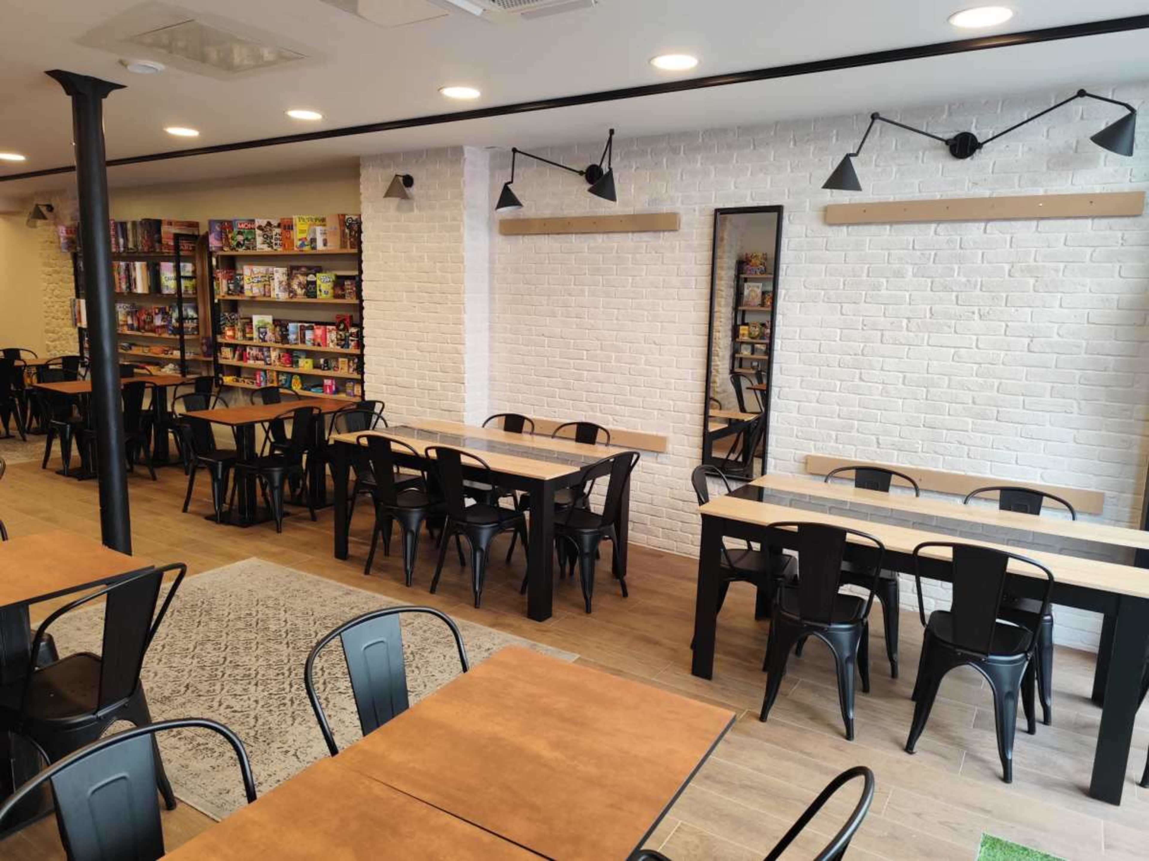 The image shows a modern interior of a café or game store with wooden tables, black metal chairs, and shelves filled with board games along one wall.