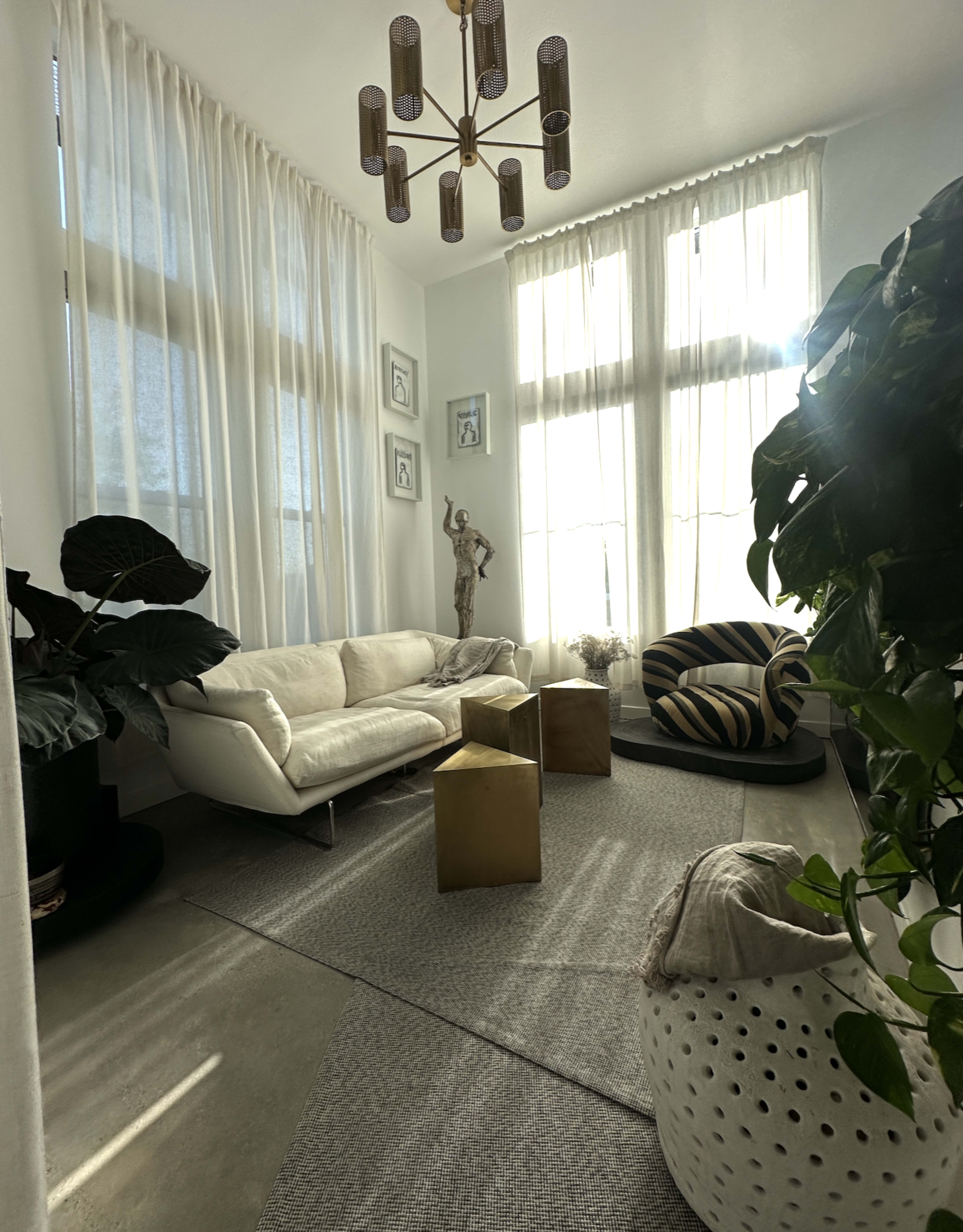 The image depicts a bright, modern living space with a white sofa, striped armchair, and large windows dressed with sheer curtains, surrounded by greenery and minimal decor.