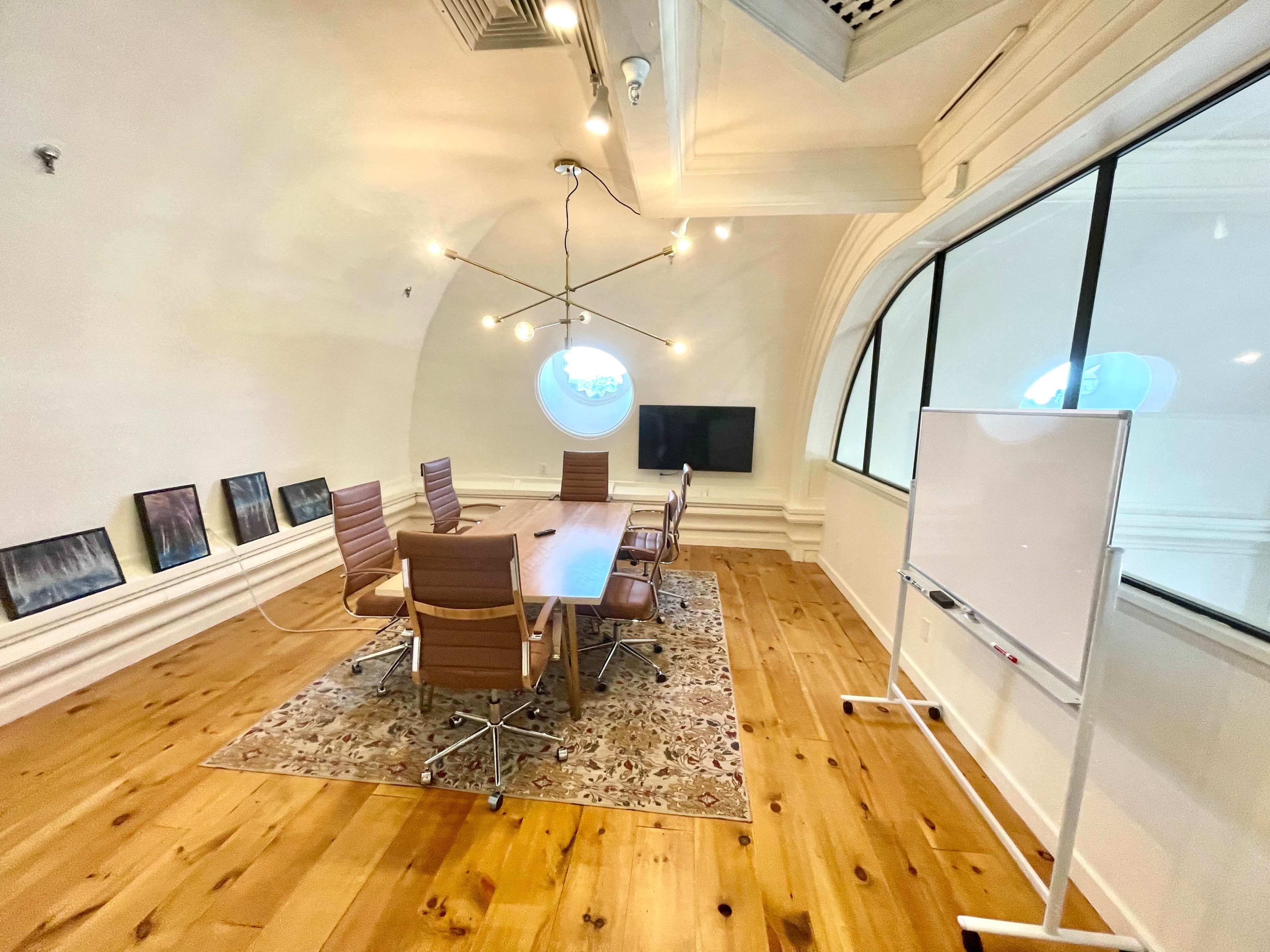 A modern conference room with a long wooden table, eight leather chairs, a whiteboard, and a large window featuring a circular design.