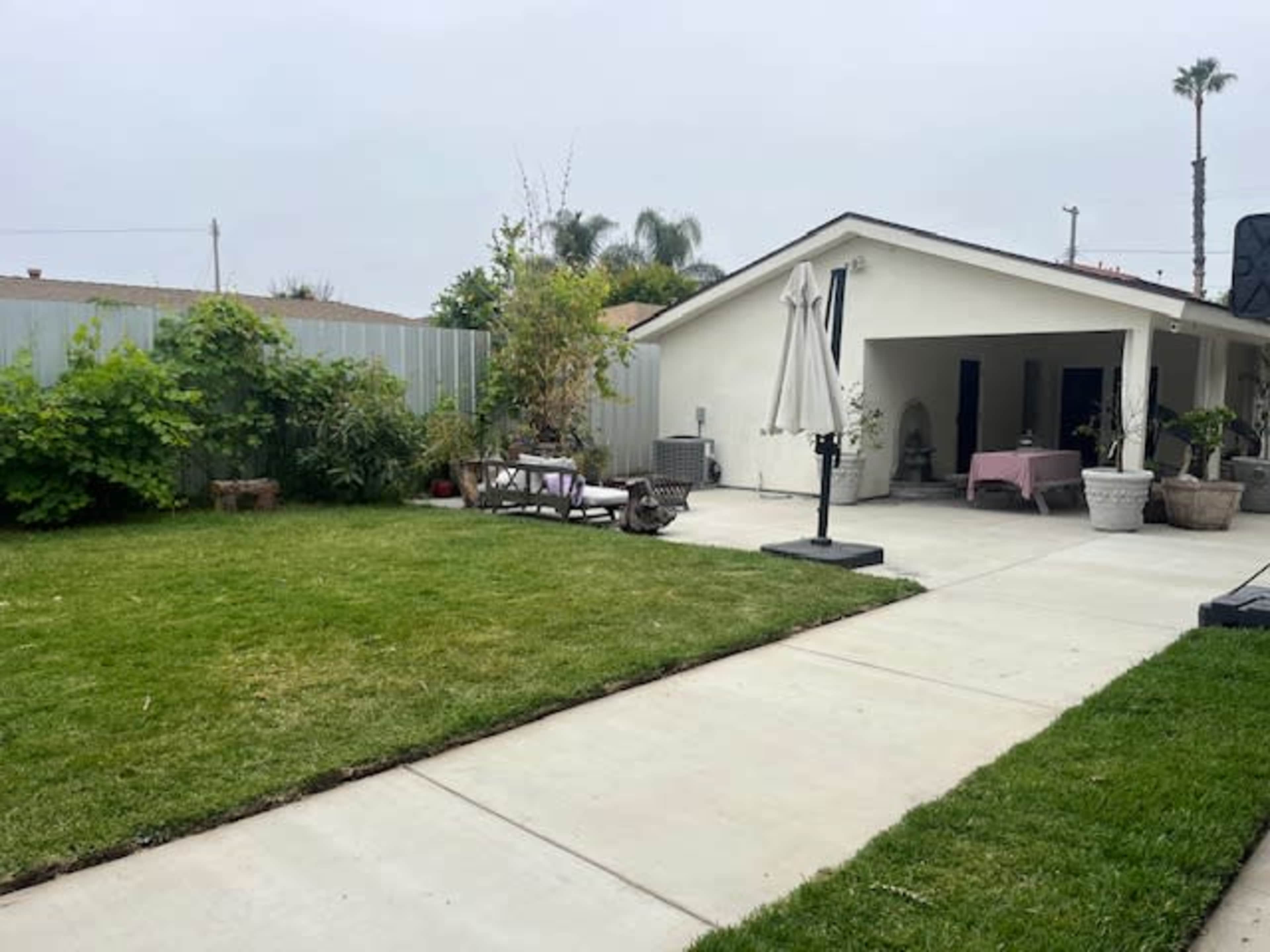 A landscaped backyard features neatly trimmed grass, a concrete pathway, outdoor seating, and a patio with a table under an umbrella.