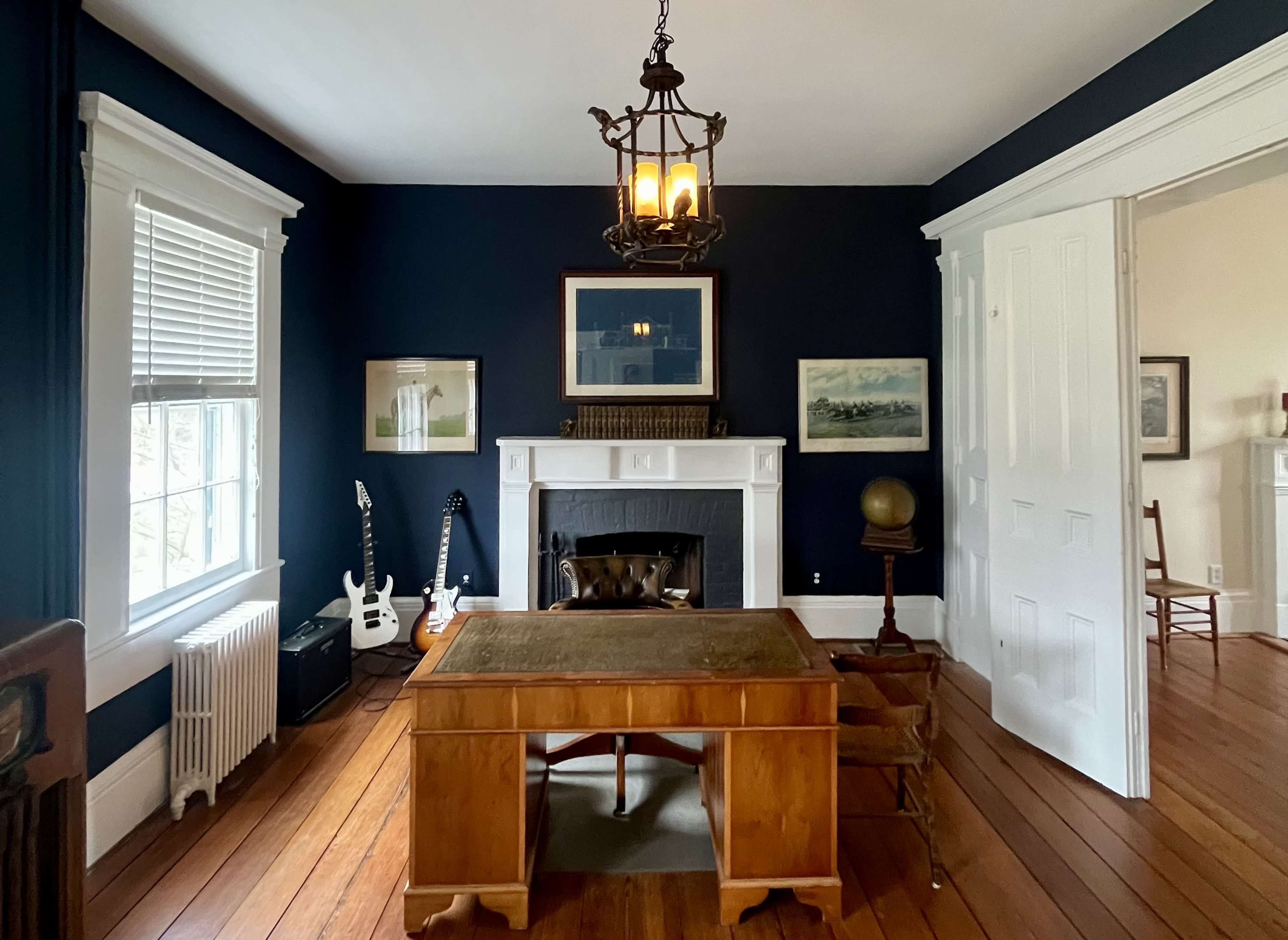 The room features a wooden desk in front of a fireplace, with dark blue walls, a chandelier, and a guitar leaning against the wall.