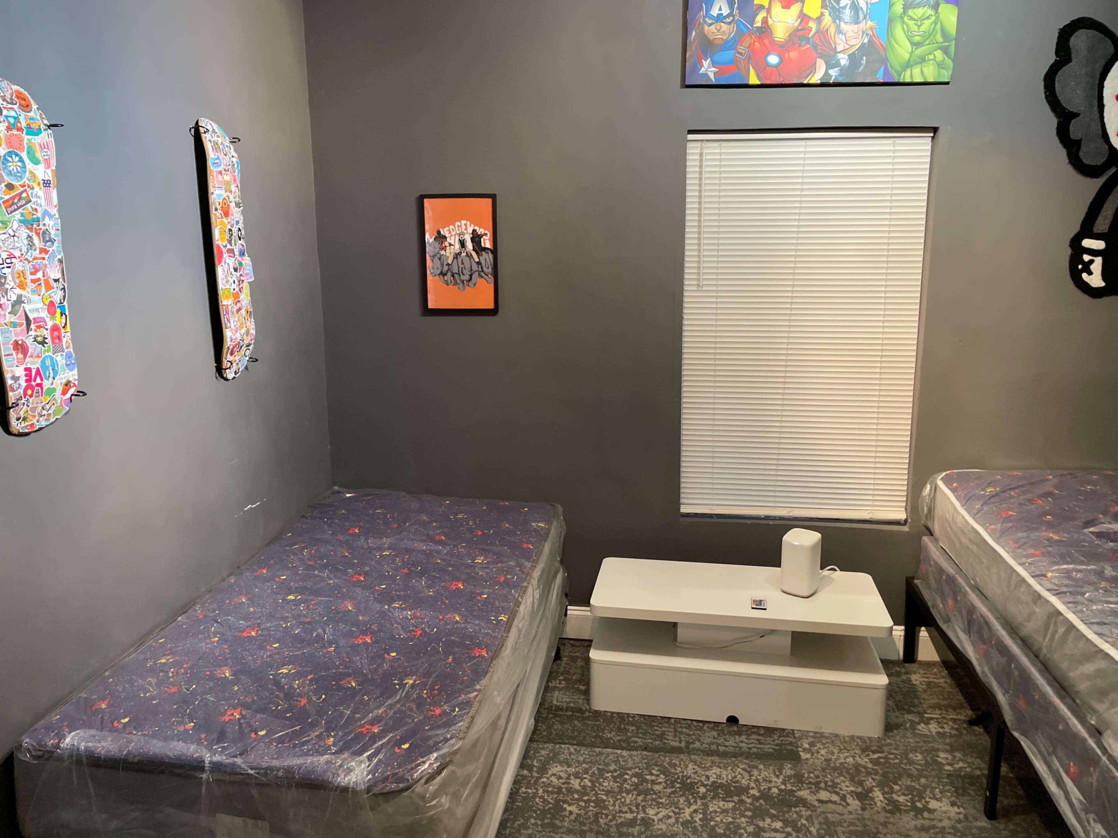 The room features two unmade beds with a central white table, a window with blinds, and colorful artwork on the walls.