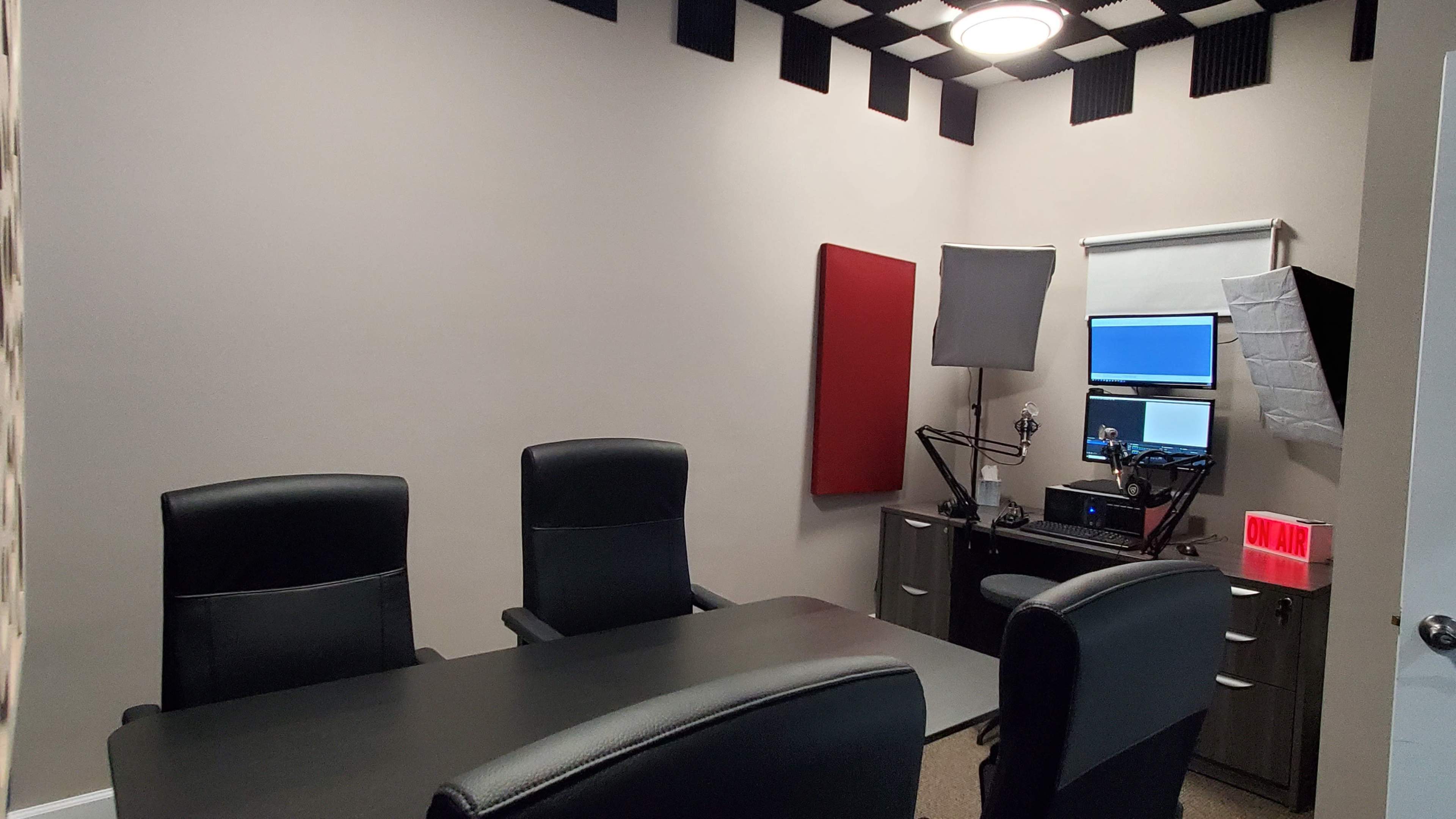 A recording studio with a long table and three black chairs, equipped with a computer, microphone, and soundproofing panels.