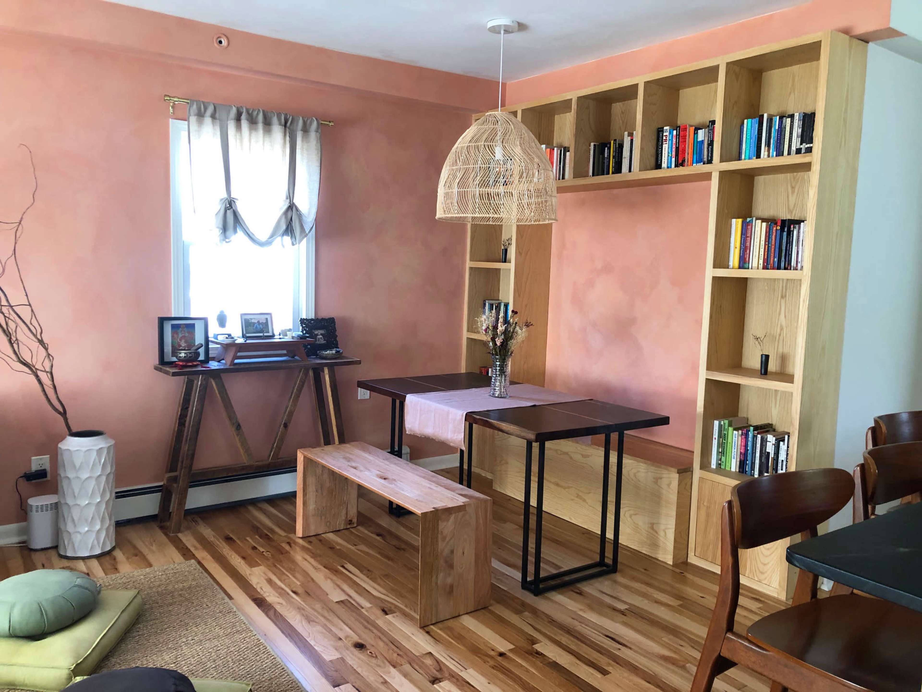 A cozy room with a wooden table and chairs, a bookshelf filled with books, and a pink wall featuring a window with sheer curtains.