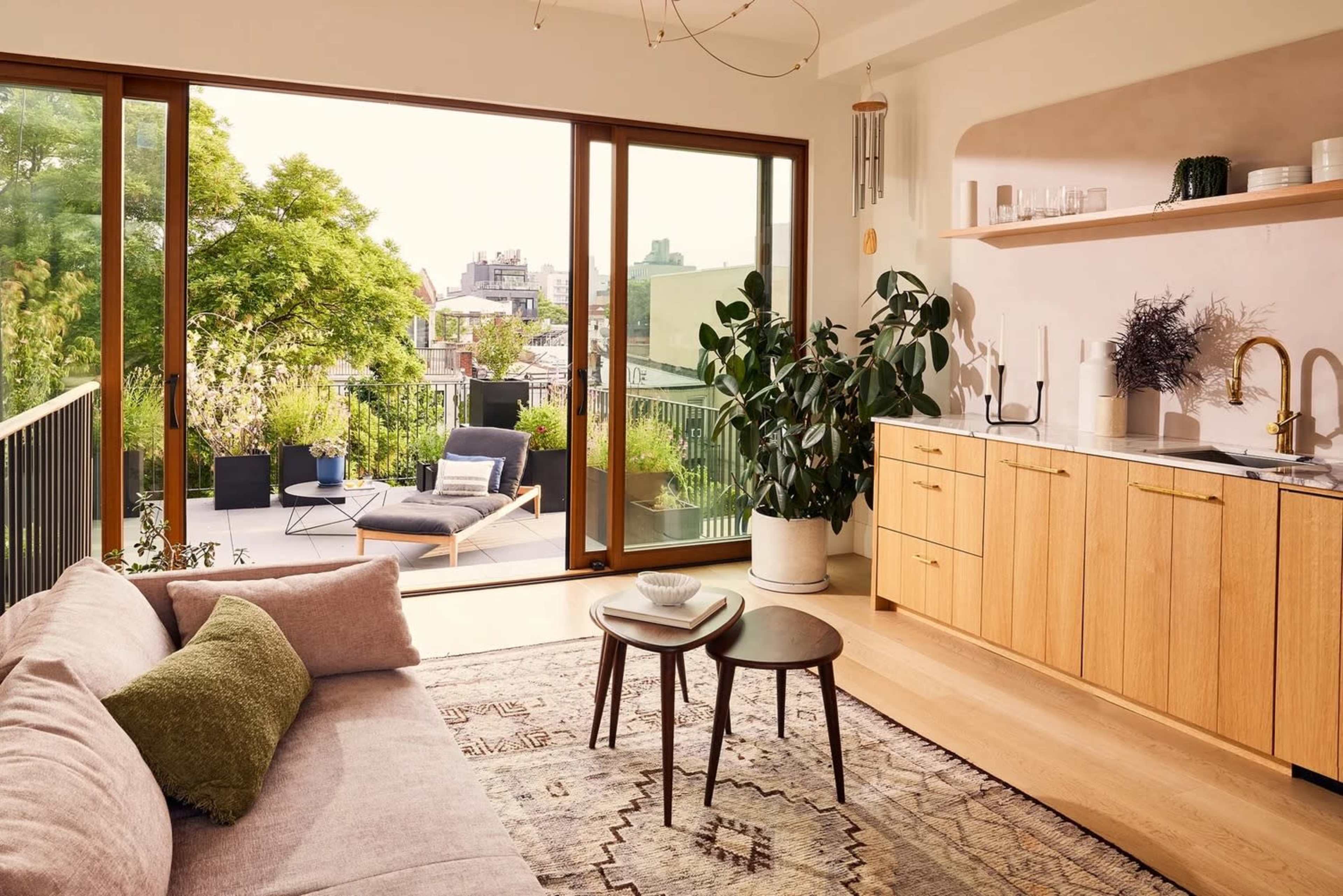 The image shows a modern living room featuring a sofa, a set of wooden tables, and a large glass door leading to a balcony with outdoor seating and greenery.