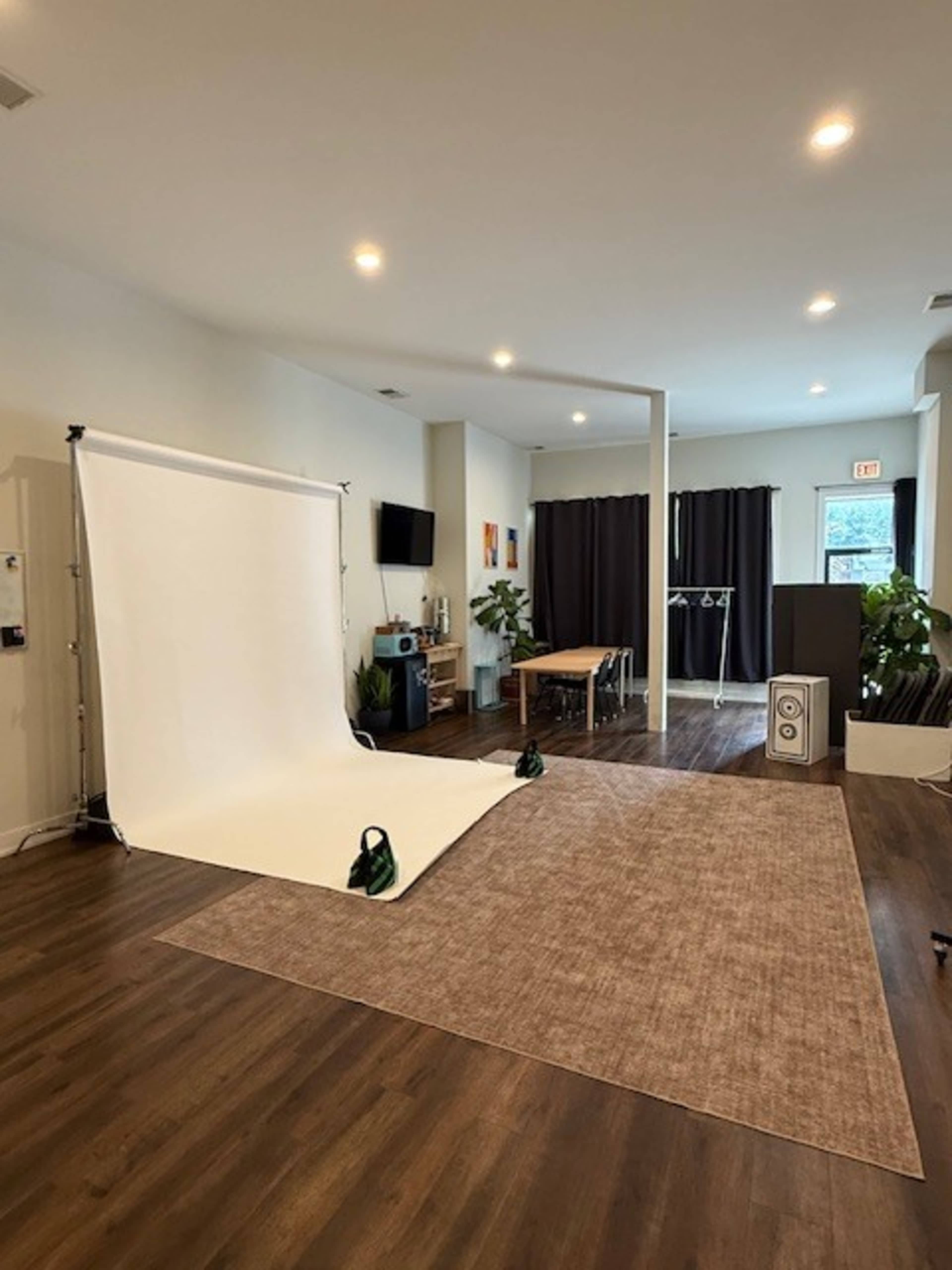 The image shows a spacious studio setup featuring a white backdrop, a wooden table, and several plants in a well-lit room.