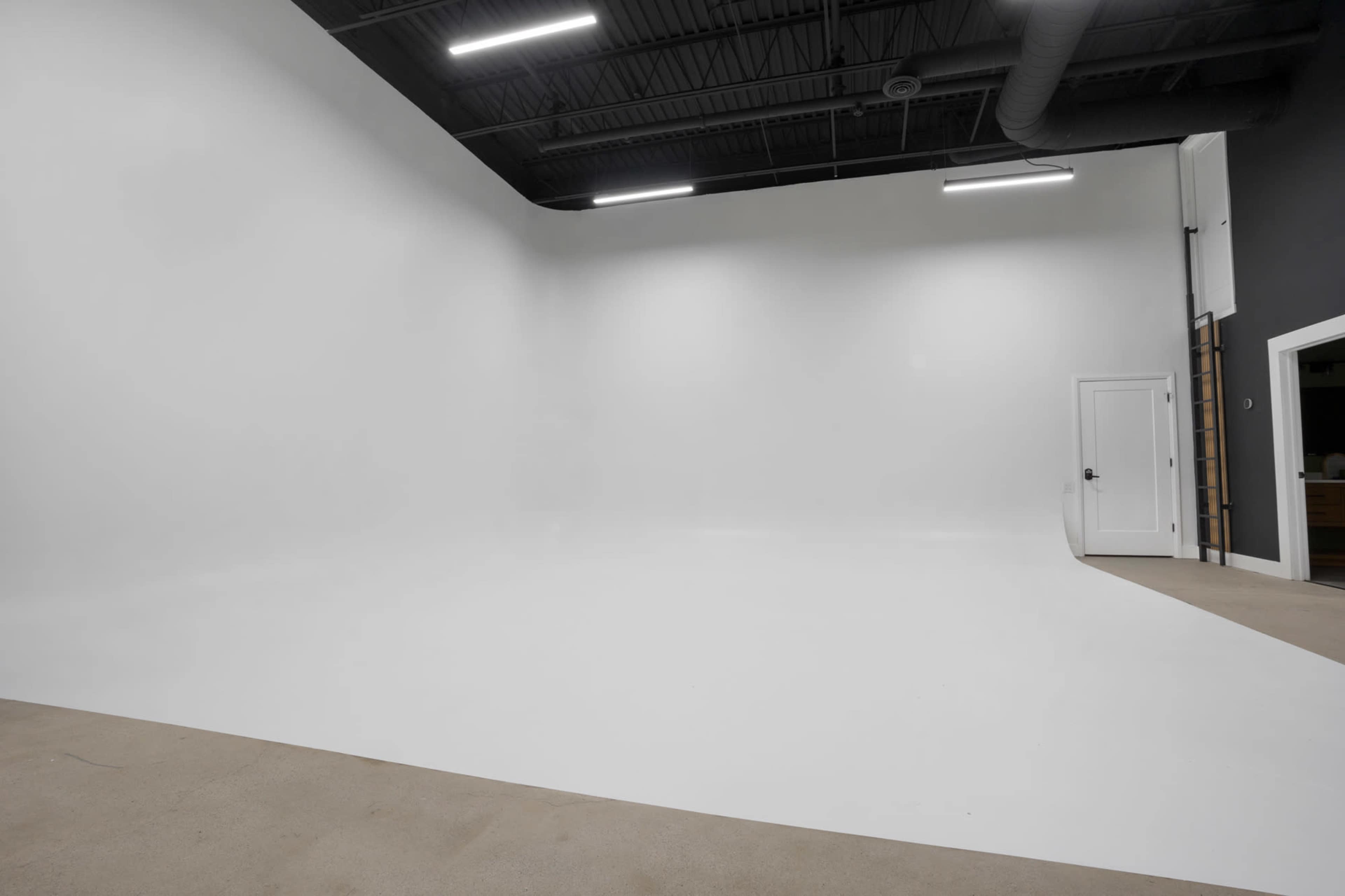 The image shows an empty white studio space with smooth walls and a light-colored floor, and a door visible on the right side.
