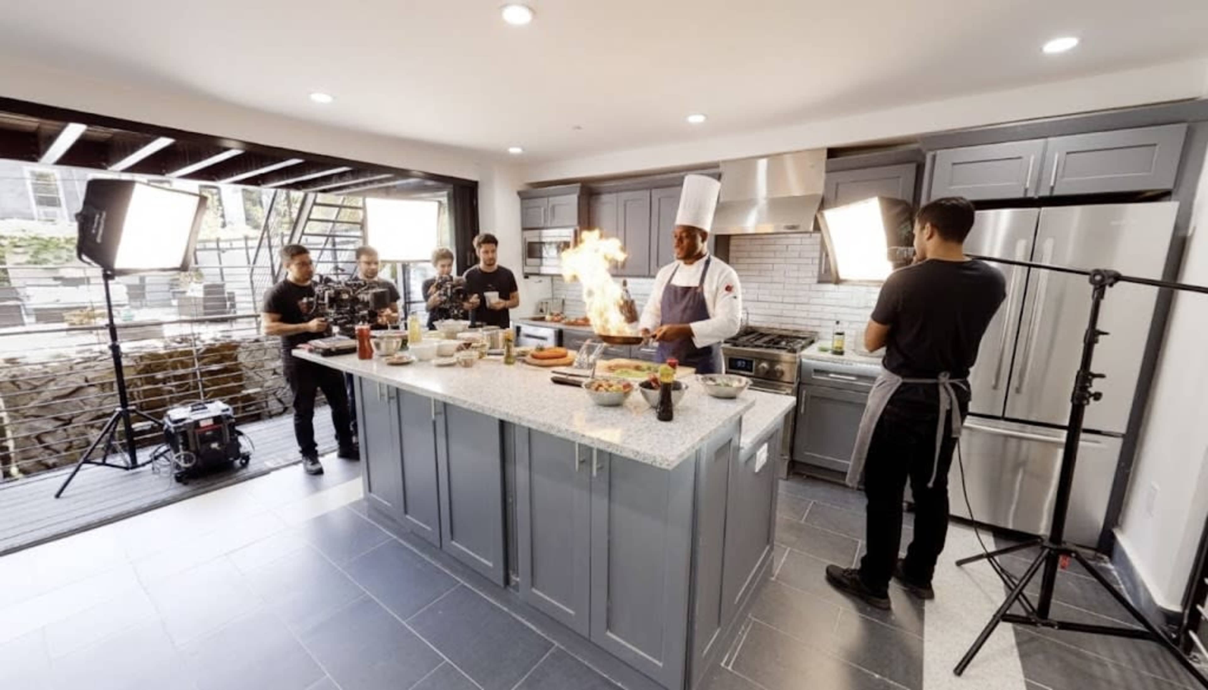 A chef in a white hat is cooking in a modern kitchen while flames rise from a dish, surrounded by a crew filming the scene.