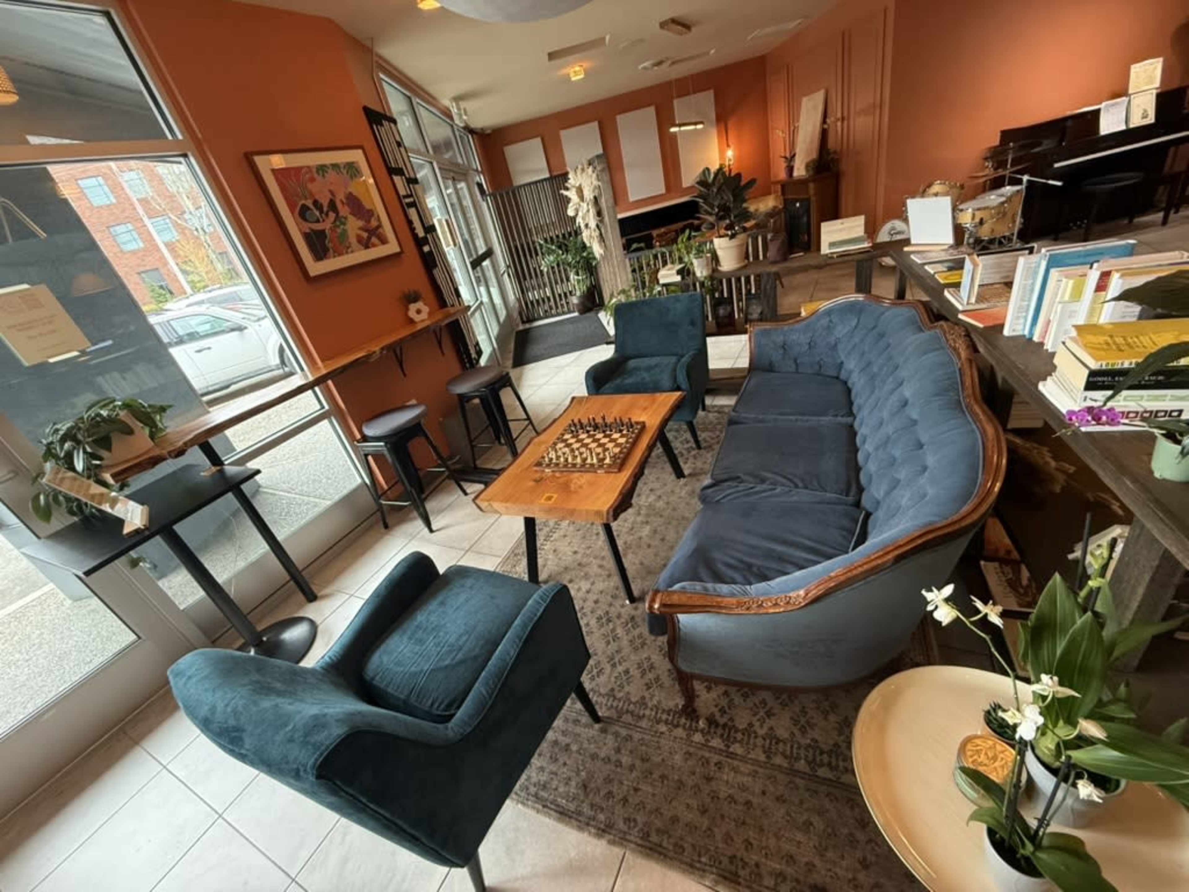 The image shows a cozy café setting with a blue sofa, two blue chairs, and a wooden table featuring a chessboard, surrounded by bookshelves and natural light.