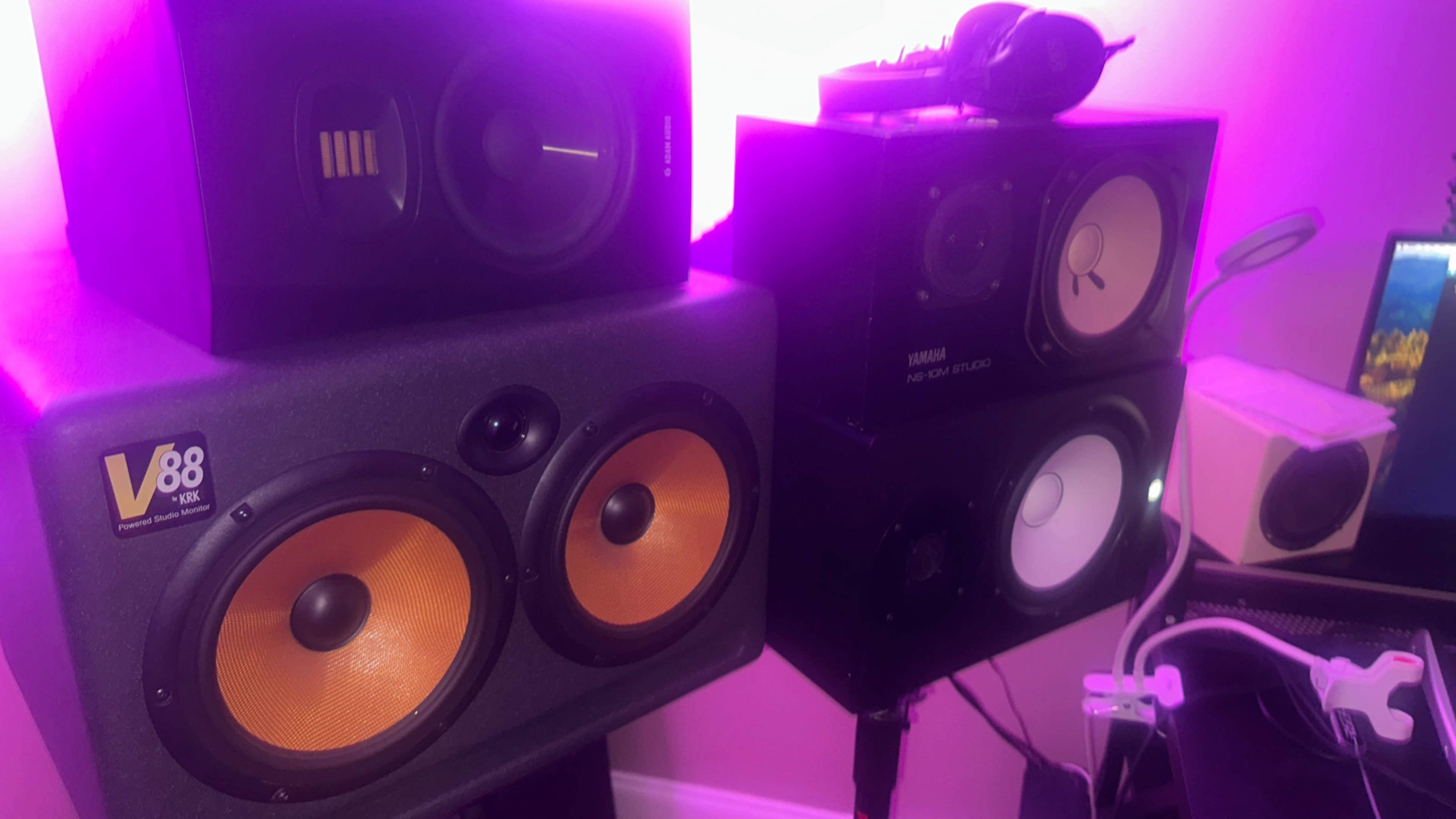 The image shows a close-up of two sets of speakers with large, orange woofers and a set of headphones placed on top of one speaker, against a pink-lit background.