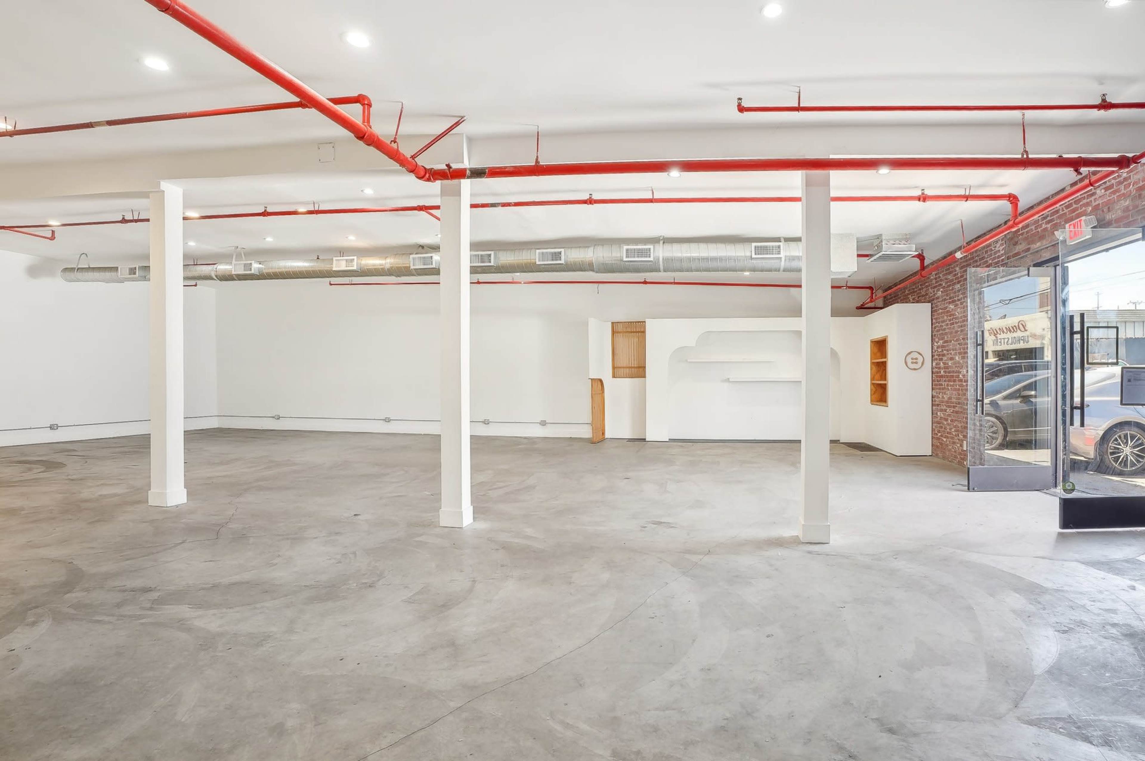 The image shows an empty commercial space with exposed red piping, concrete floors, and large glass doors leading to the exterior.