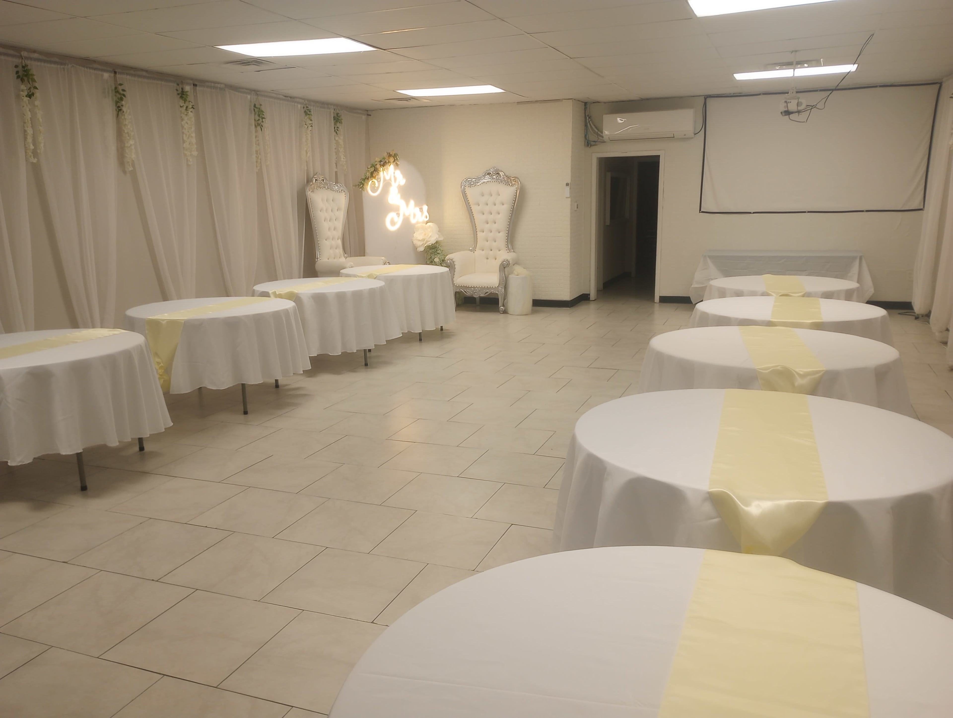 The image shows a spacious event hall with several round tables covered in white tablecloths and a yellow runner, flanked by decorated walls and chairs at one end.