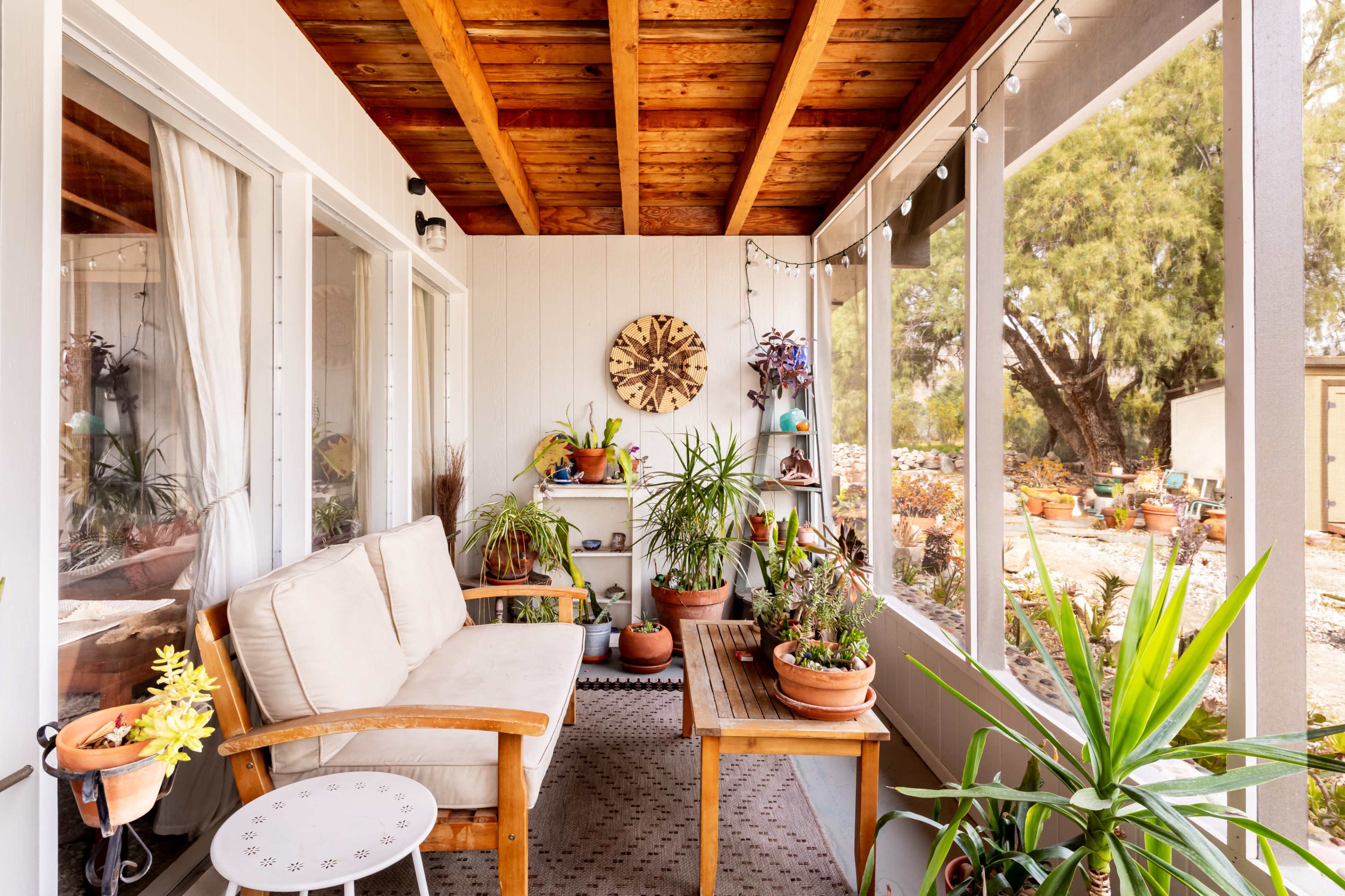 The image shows a covered porch with wooden beams, a seating area, and various potted plants arranged on tables and the floor.