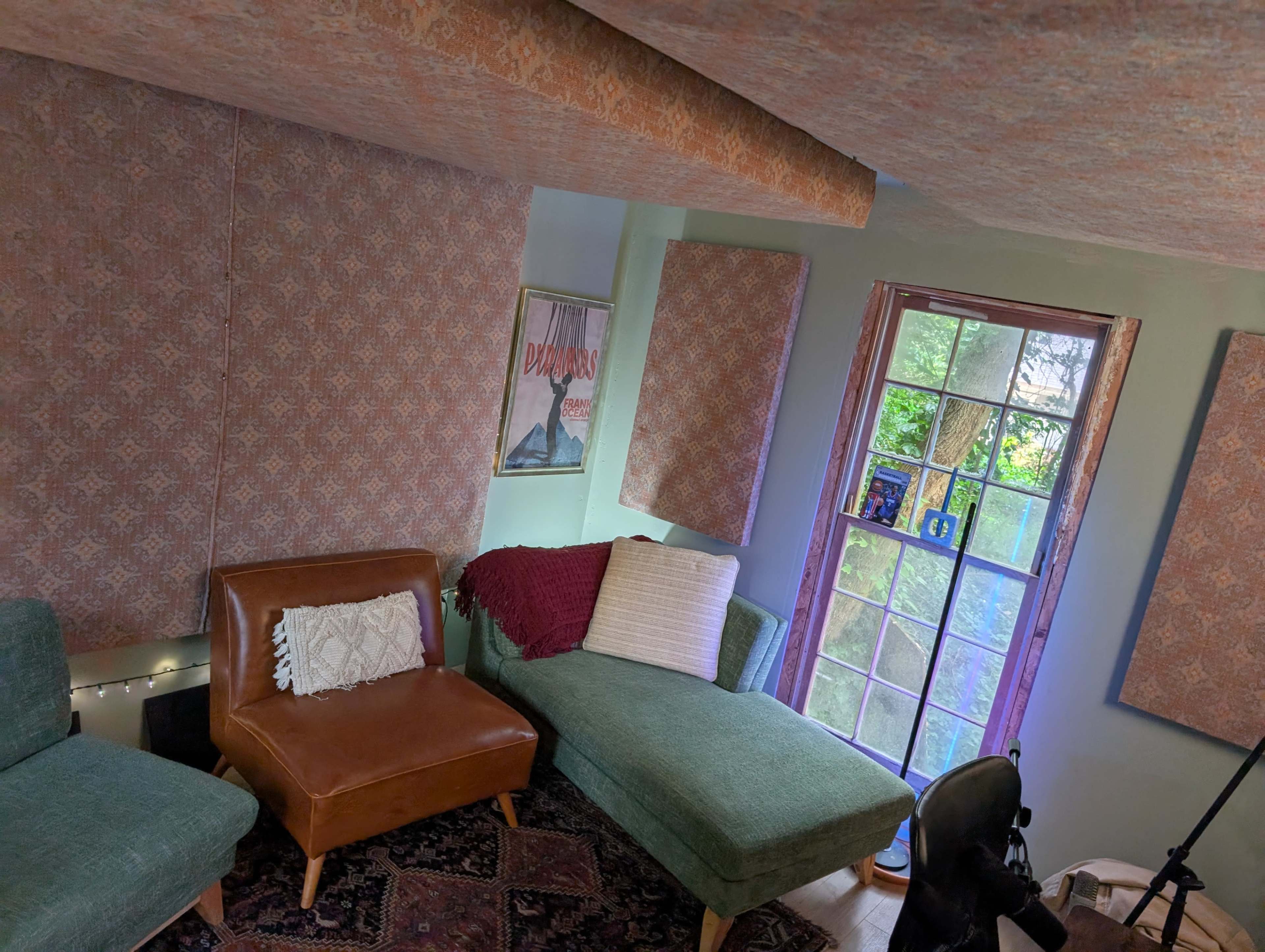 The image shows a cozy room with a brown chair and a green sectional couch, decorated with patterned wall panels and a window overlooking greenery.