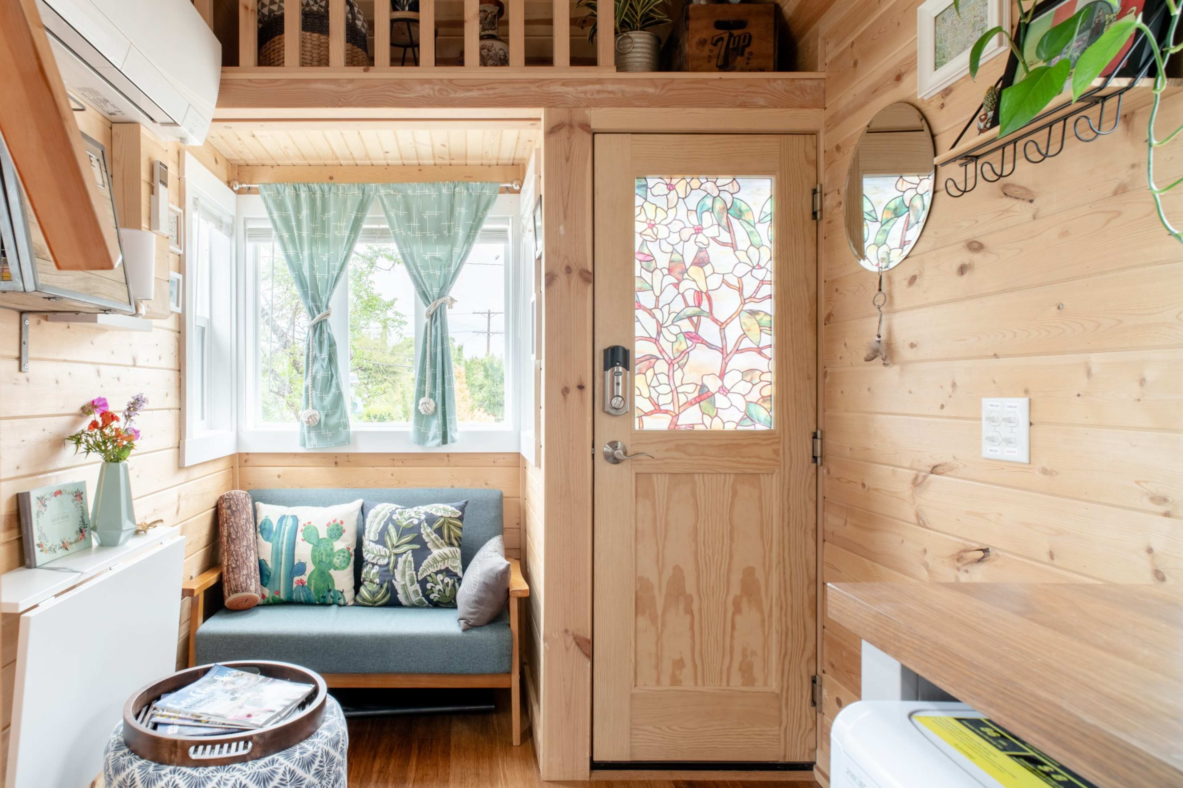A small, wooden interior space featuring a couch, a decorative door with stained glass, and a window with green curtains.