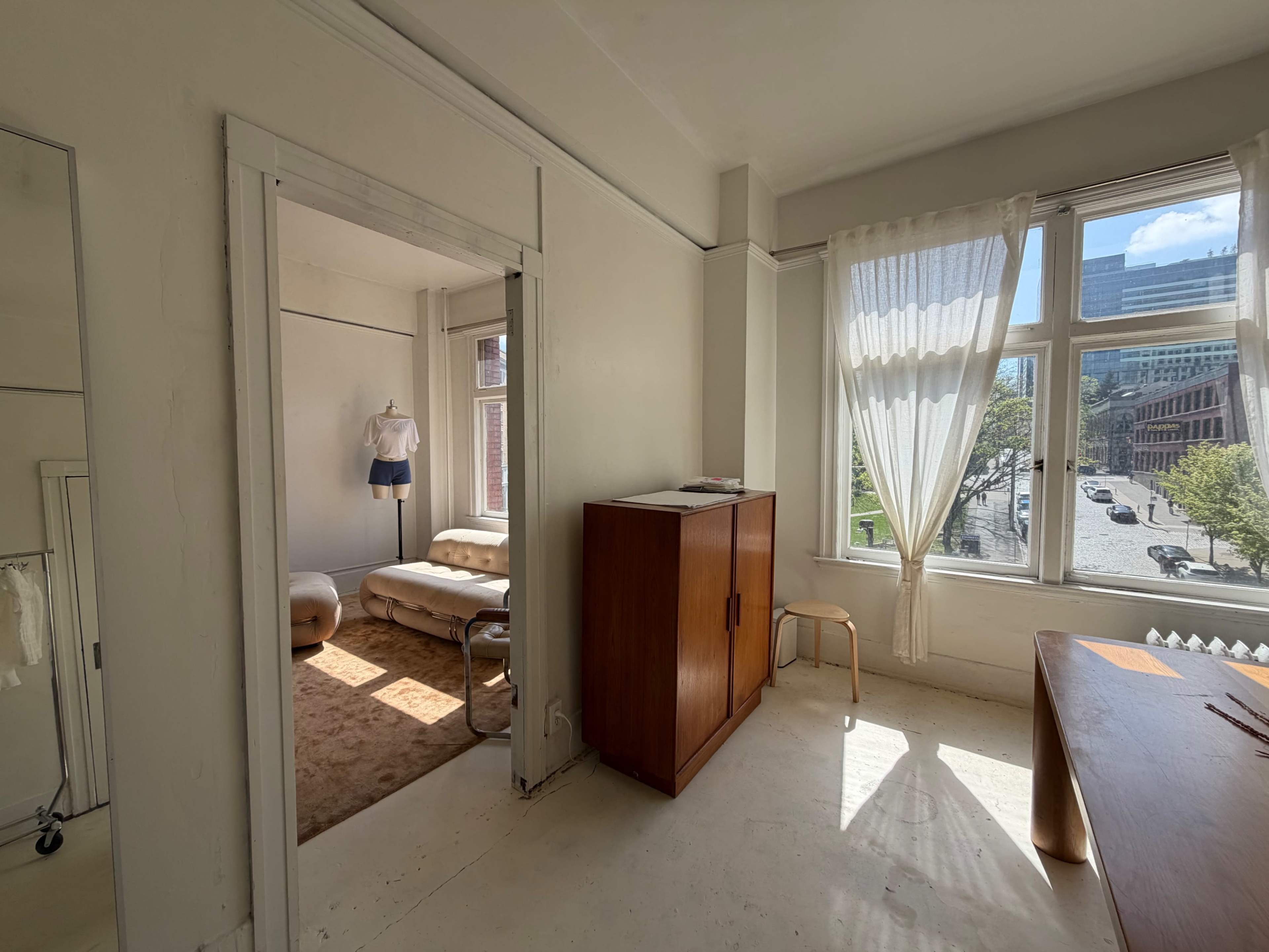 The image shows a bright, minimalist room with a light-colored floor, a wooden cabinet, and a view of a street through a large window.