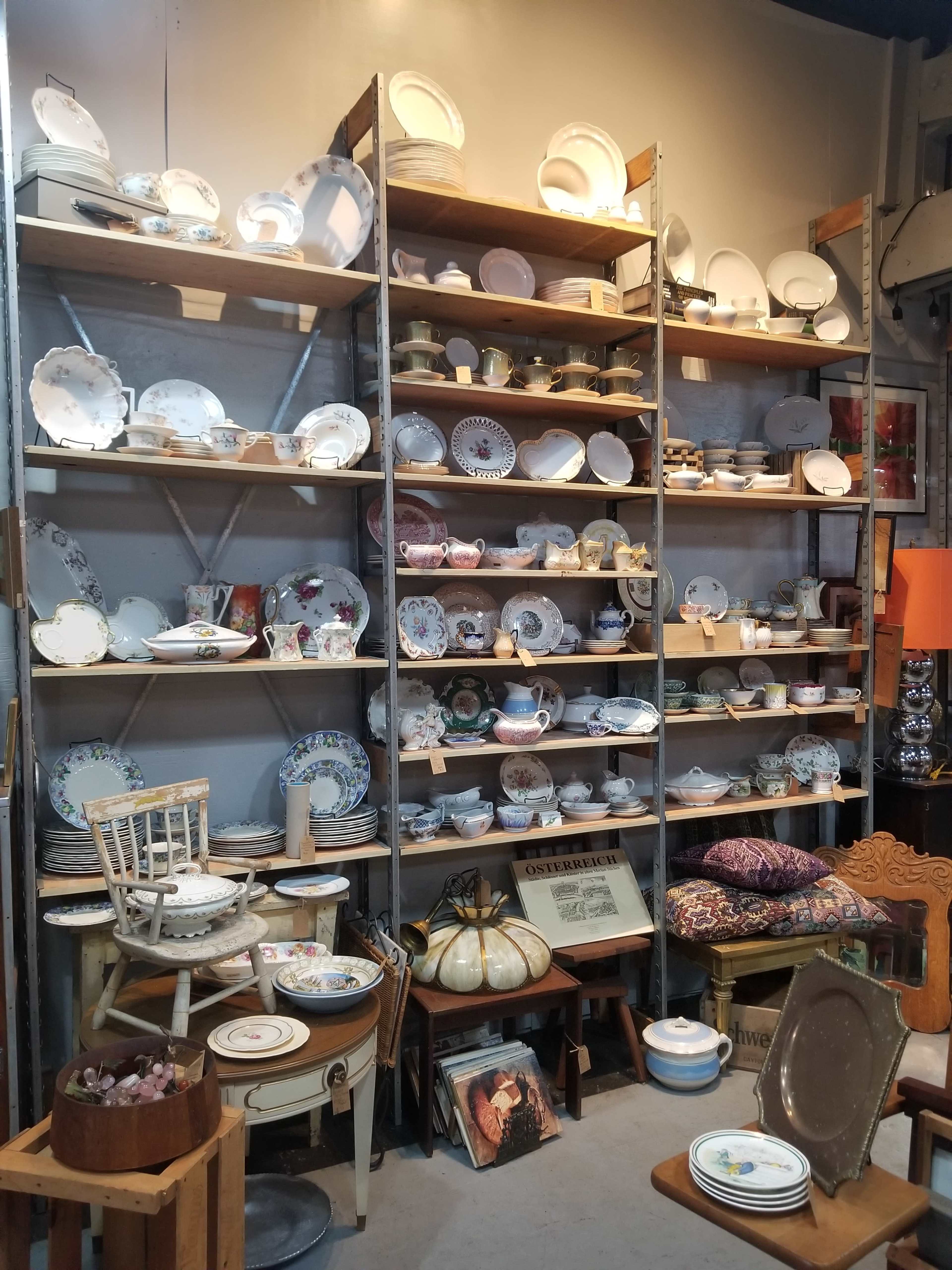 A tall shelving unit displays a collection of various plates, bowls, and decorative items alongside furniture and cushions in a store.