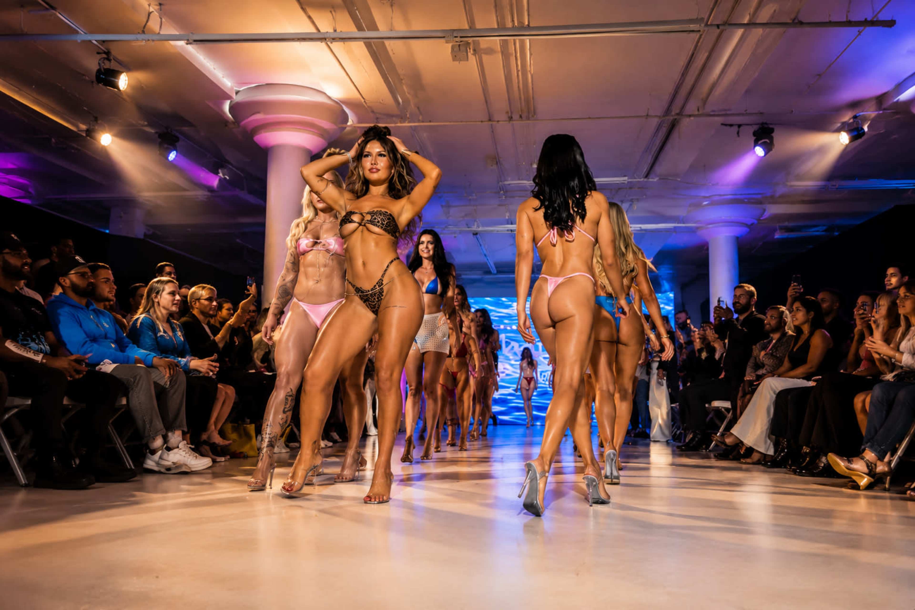 Models walk down a runway during a fashion show, showcasing swimwear amidst an audience of spectators.