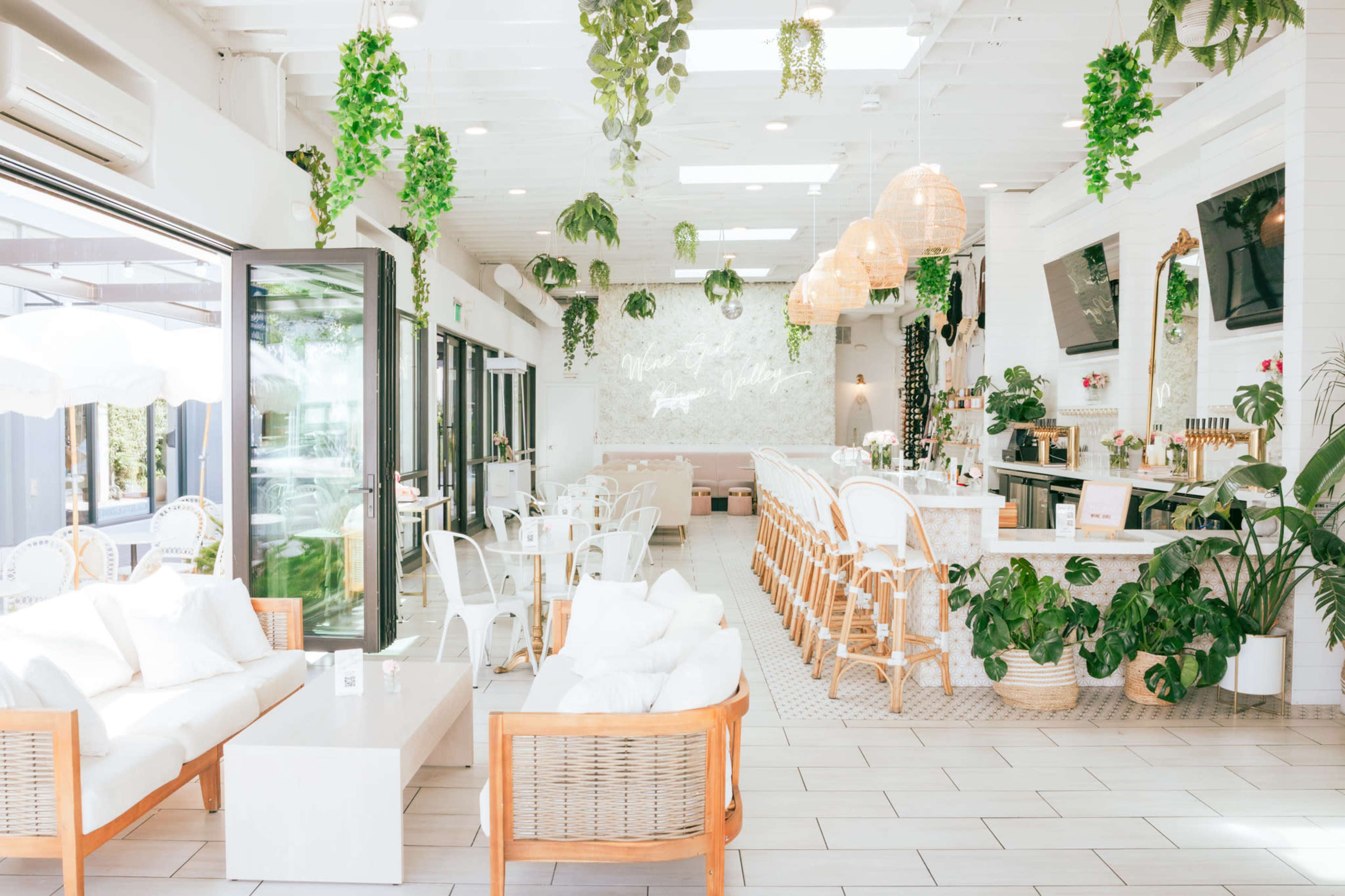 The image shows a modern café interior with white walls, rattan furniture, and abundant greenery hanging from the ceiling.