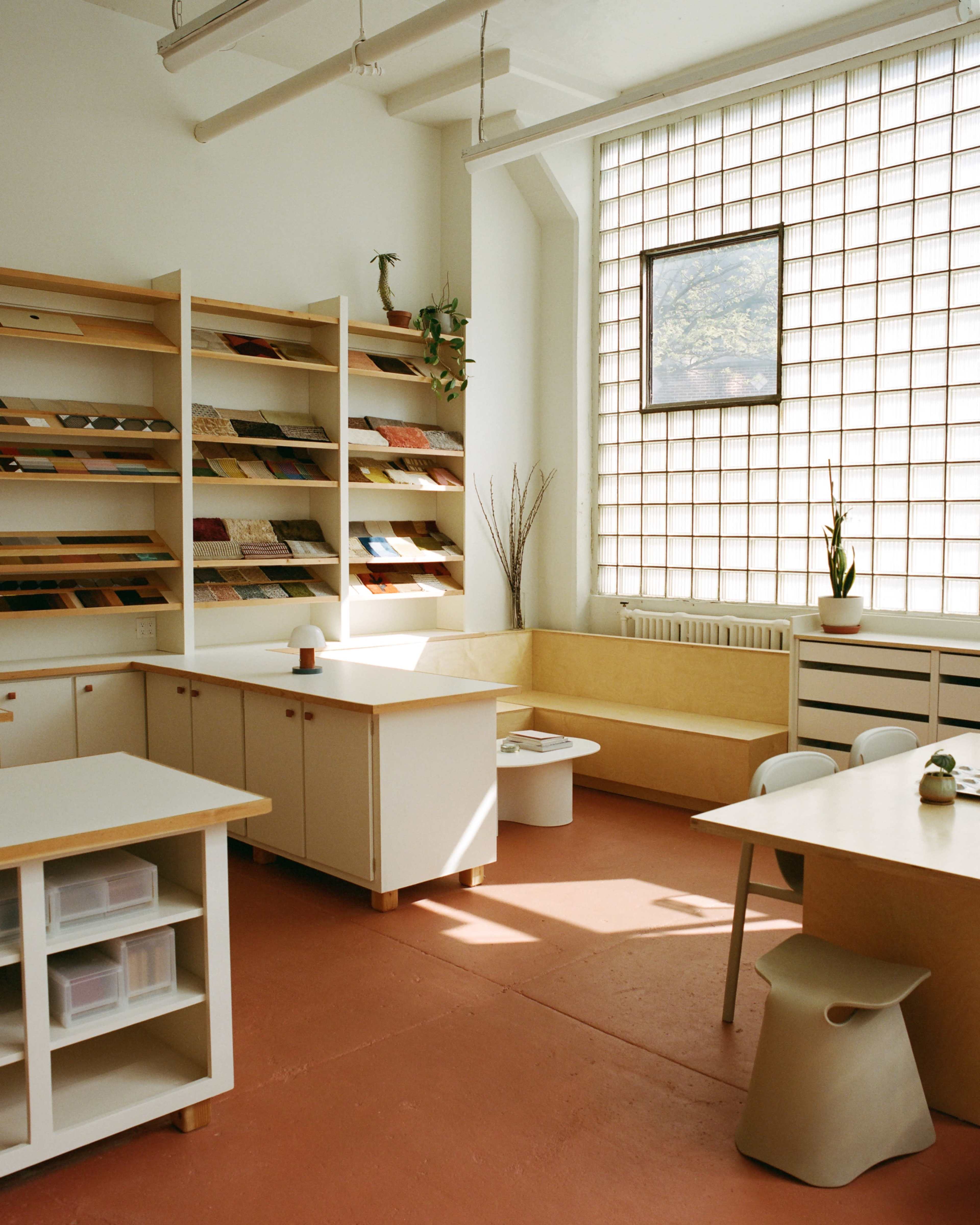The image shows a bright, minimalistic office space with wooden shelving displaying samples, a large window, and a seating area.