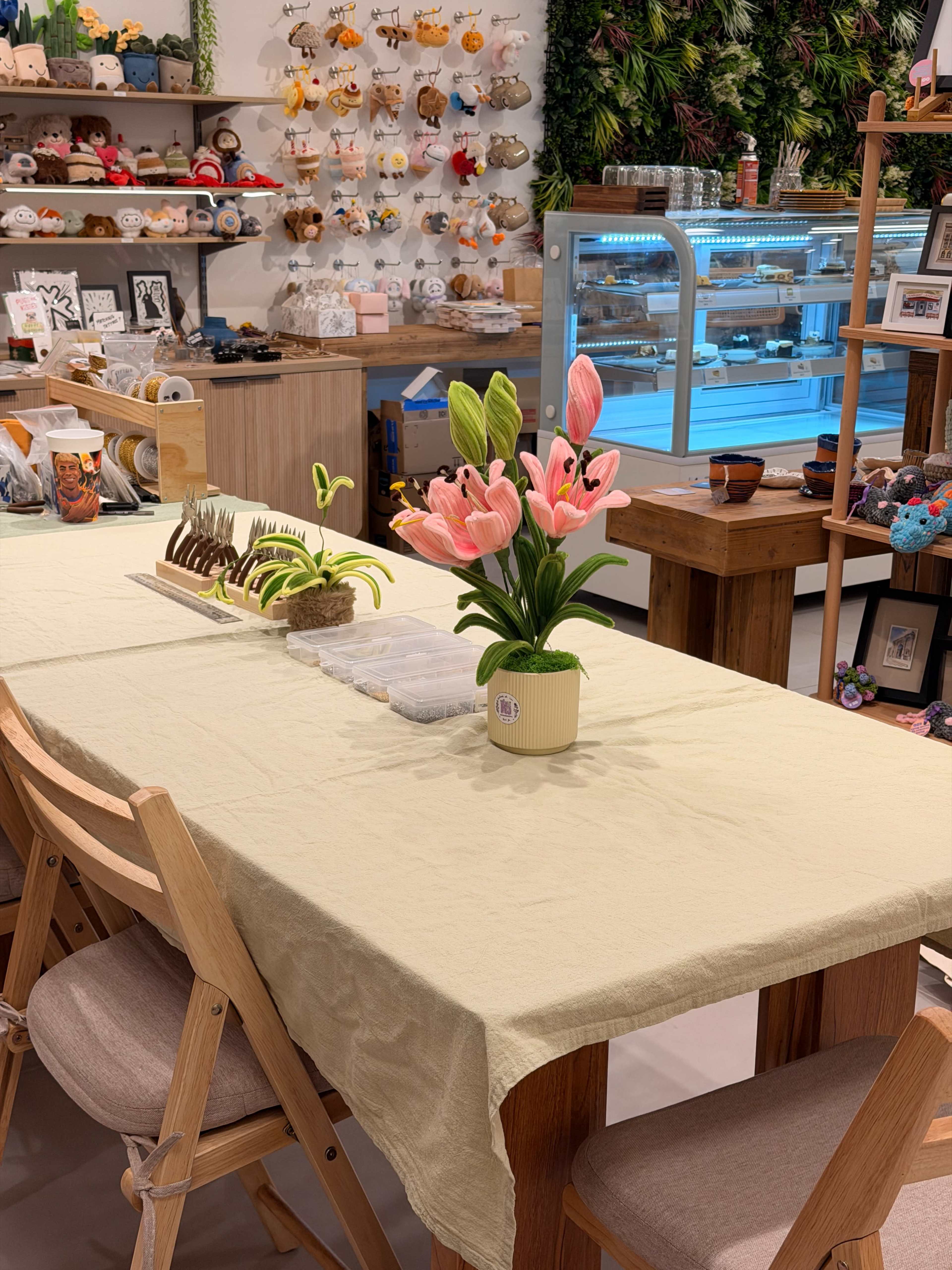 A table is set with a light-colored tablecloth and a vase of pink flowers, surrounded by wooden chairs in a shop displaying various handmade crafts and decorations.