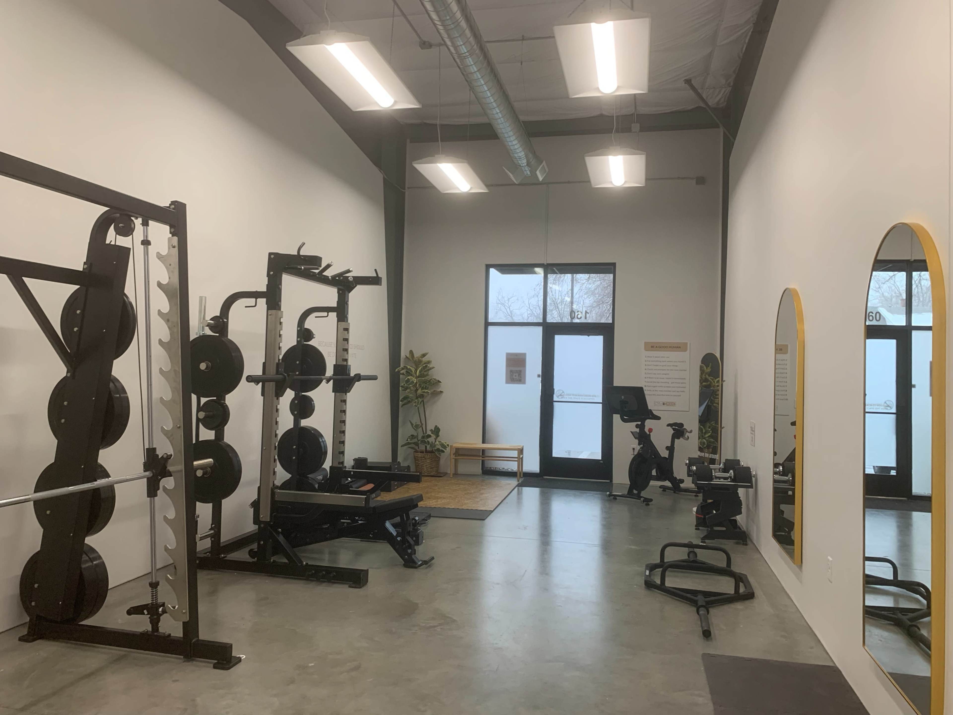 The image shows a modern gym interior featuring a weight rack, bench, exercise bike, and large mirrors, with large windows allowing natural light.