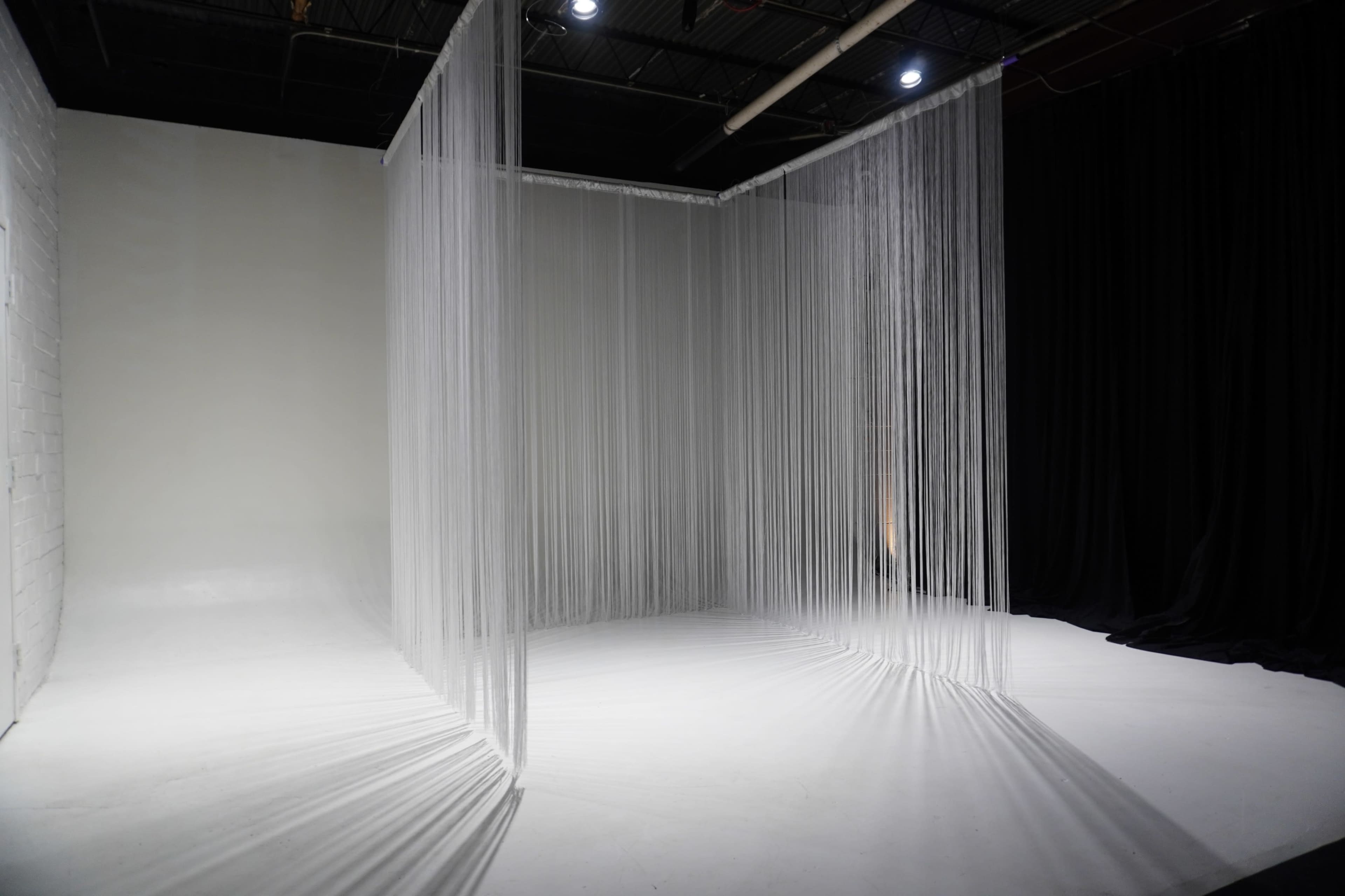 The image shows a minimalist studio space divided by thin, vertical strands of material, with a contrasting black backdrop on one side.