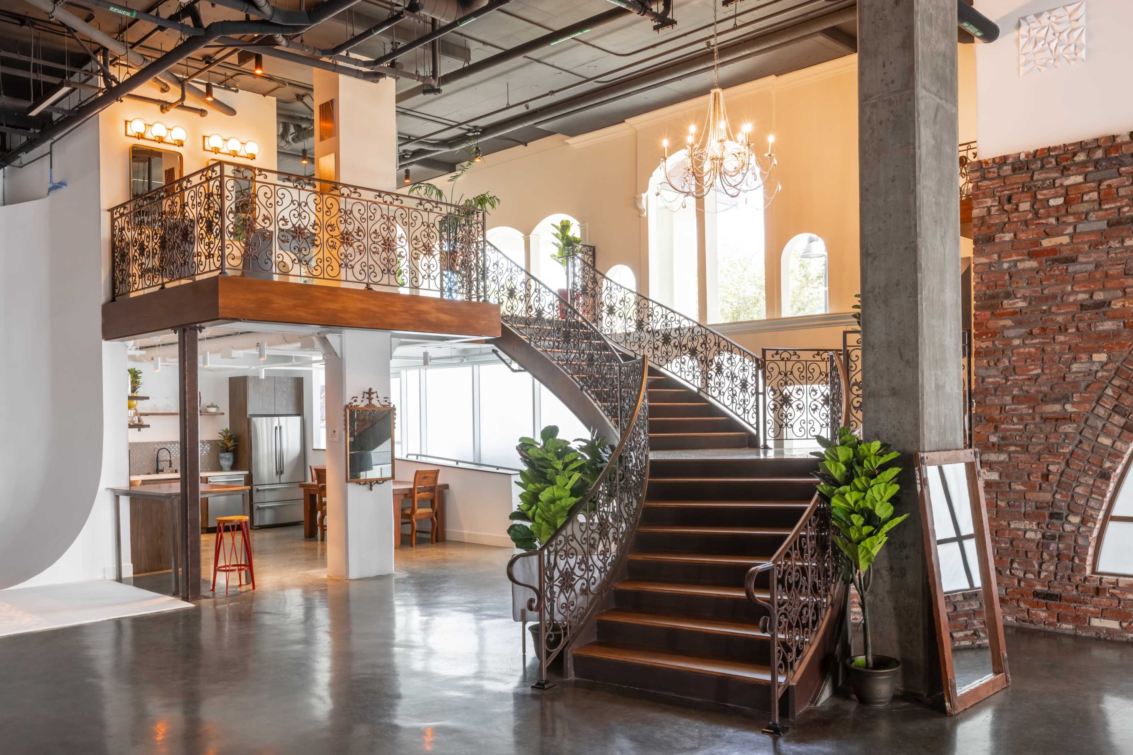 A stylish interior of a spacious hall with a grand staircase, intricate wrought iron railings, large windows, and exposed brick walls.
