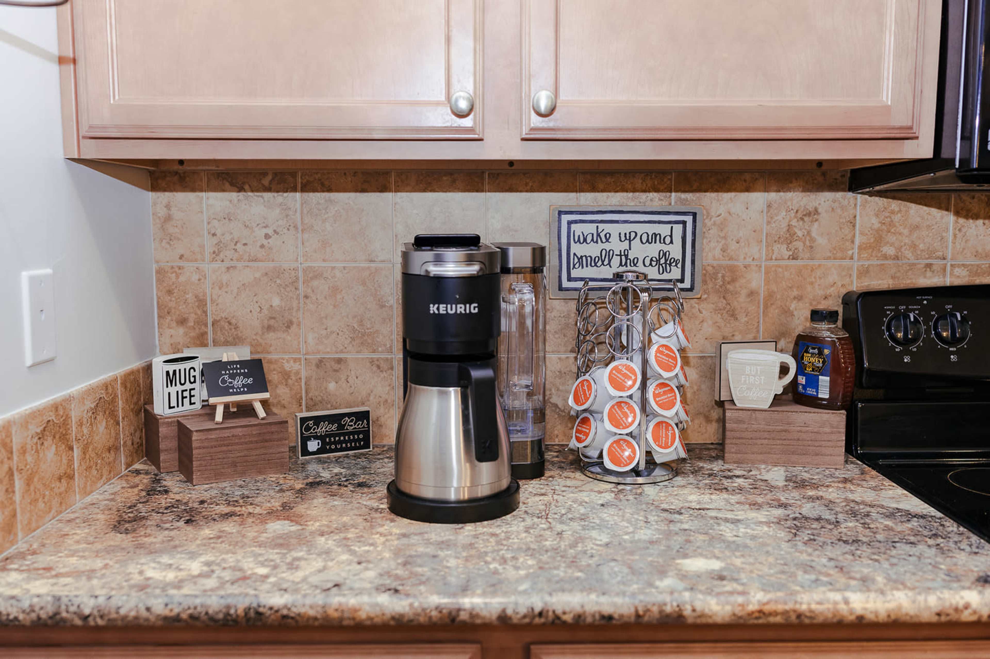 A coffee station features a Keurig coffee maker, decorative signs, and various coffee-related items arranged on a countertop with a tiled backsplash.