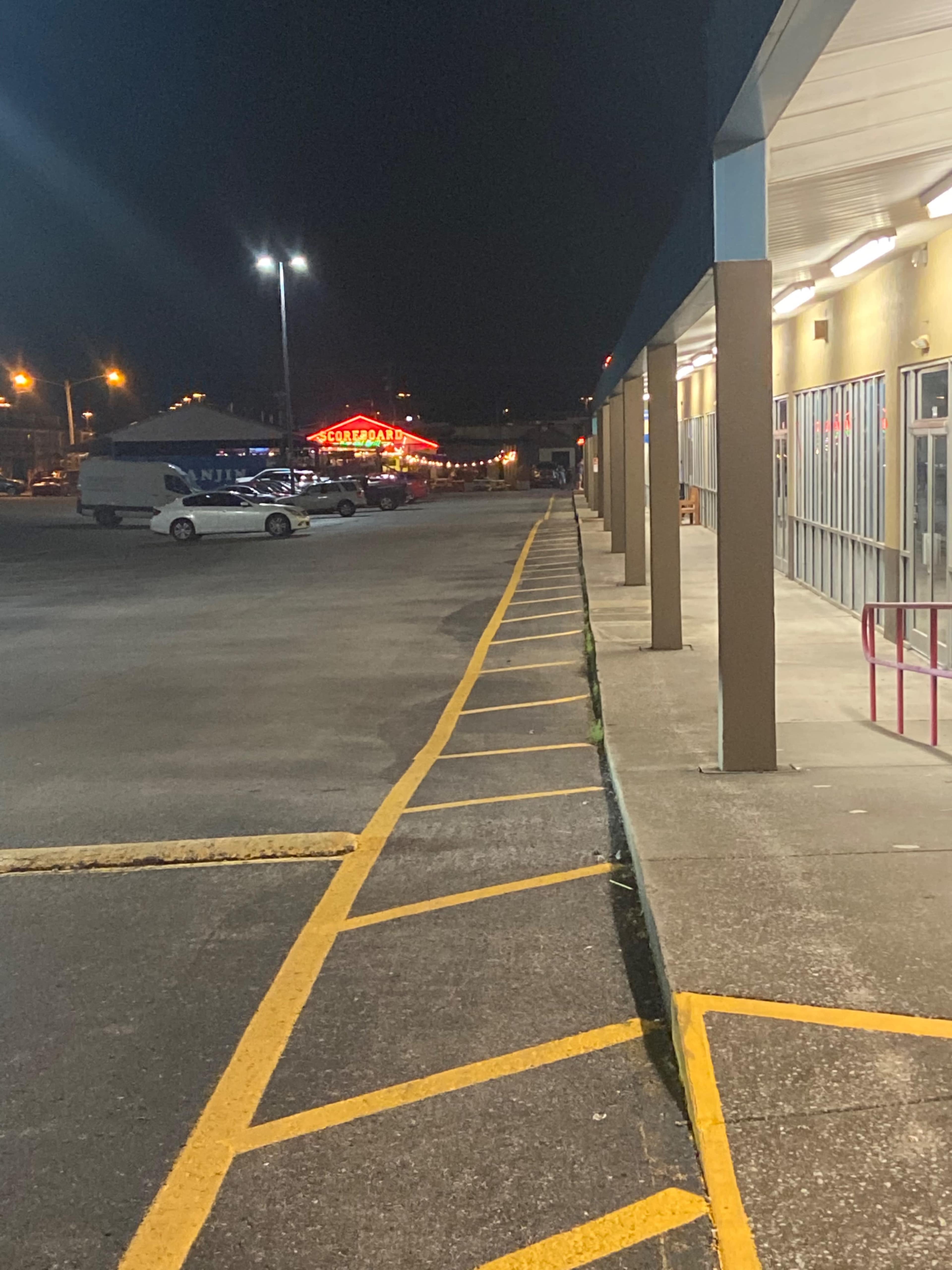 A dimly lit parking lot extends alongside a row of storefronts, with a bright neon sign visible in the distance.