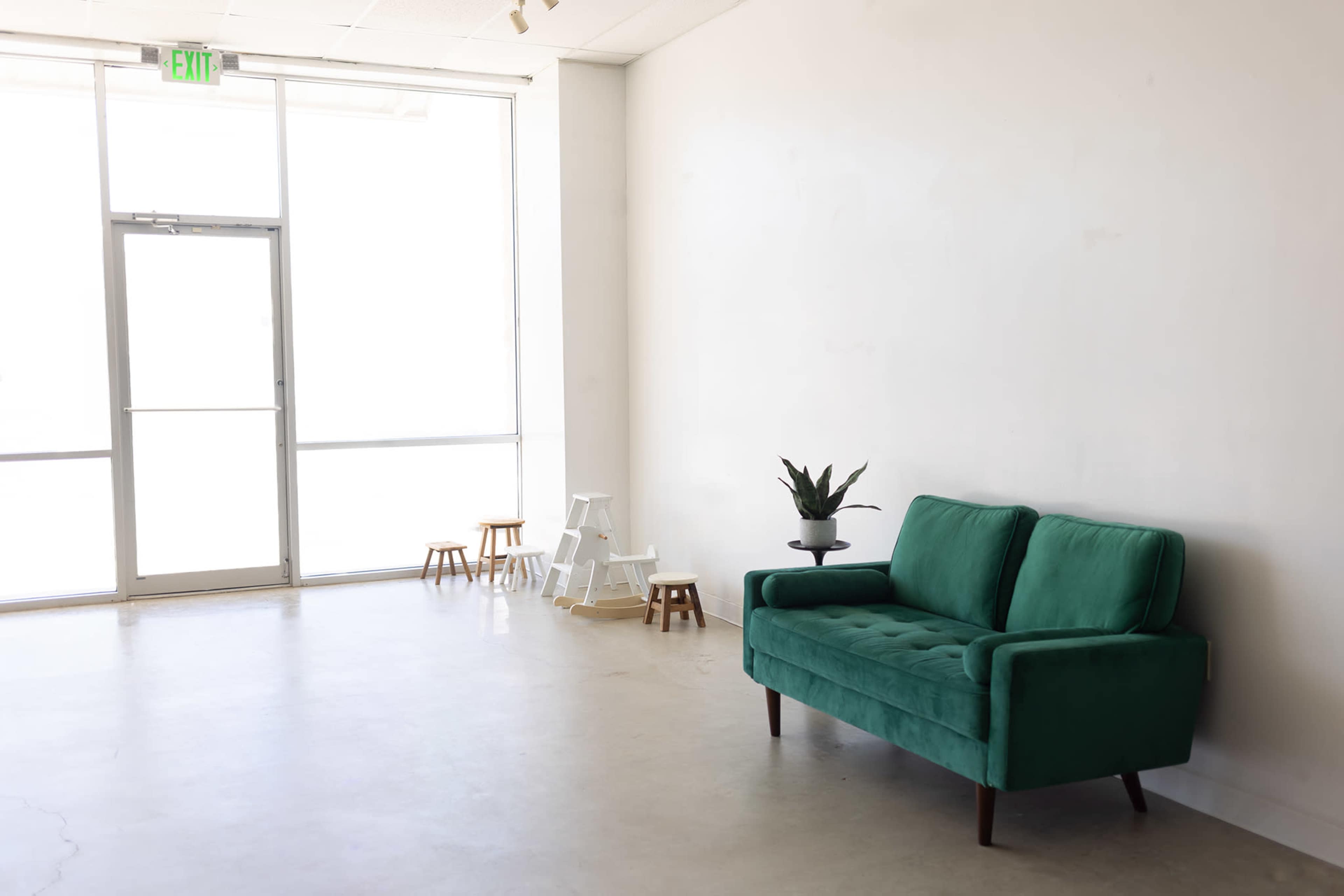 The image features a minimalist room with a green couch, a small plant on a table, and a few wooden stools near a large window.
