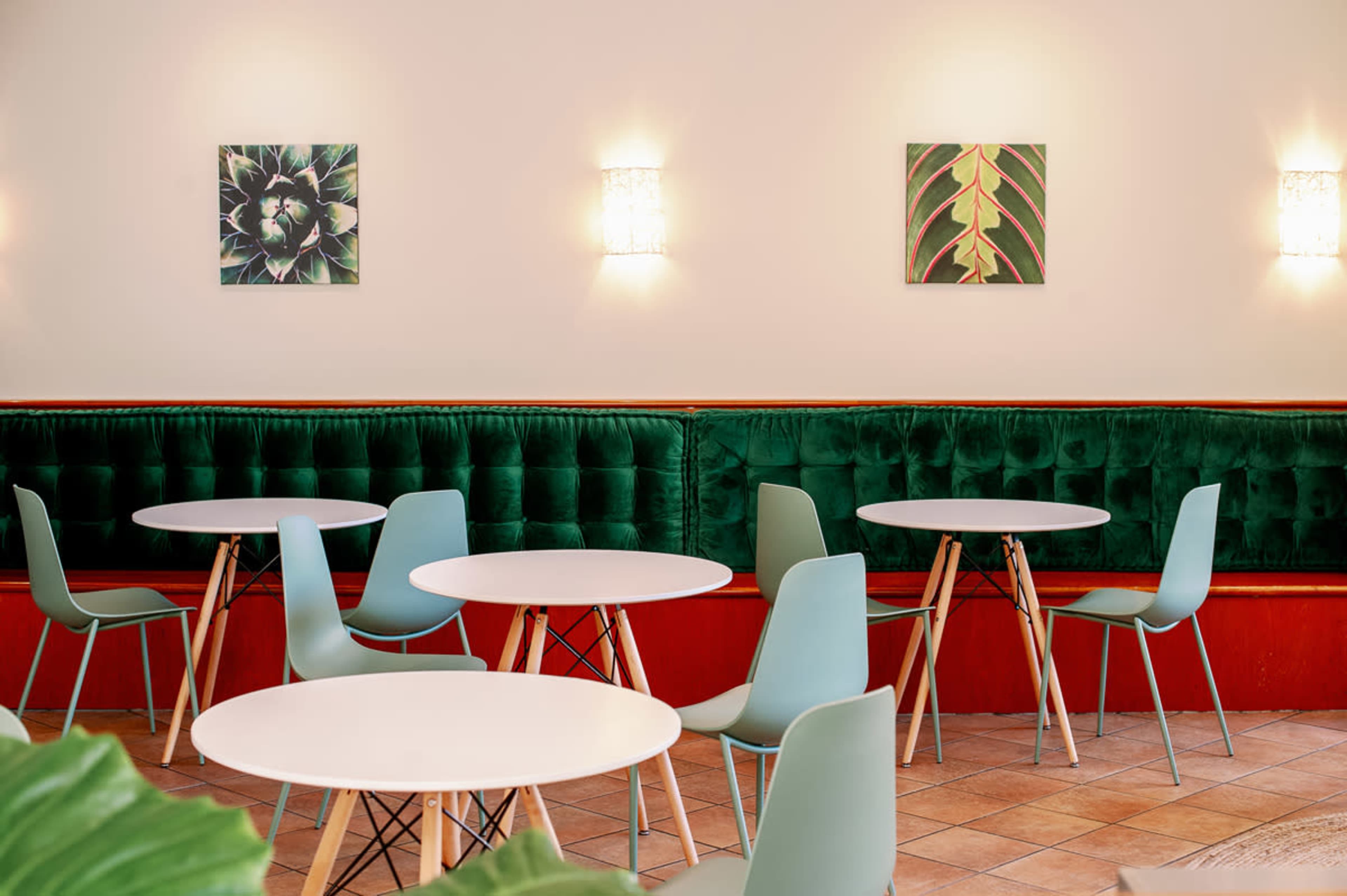 A cafe interior featuring round tables, mint green chairs, and green velvet seating, with botanical artwork on the walls.