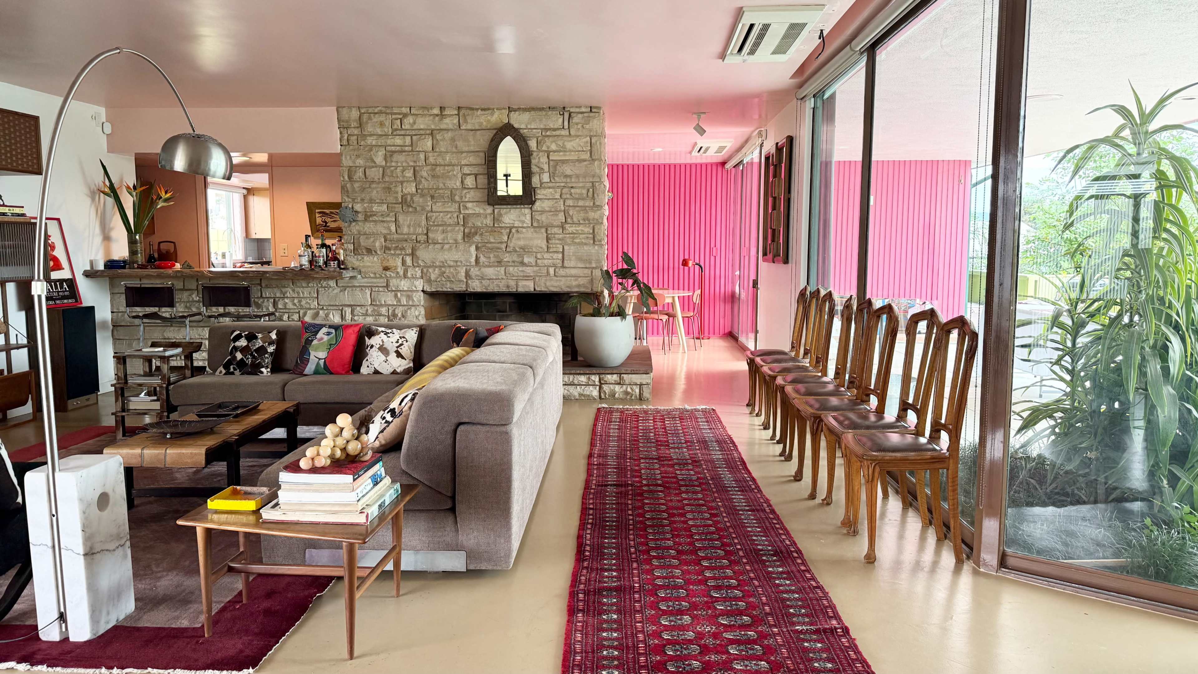 The scene depicts a modern, open living space with a stone fireplace, a large sofa, and a row of wooden chairs along one side, accented by colorful decor and plants.