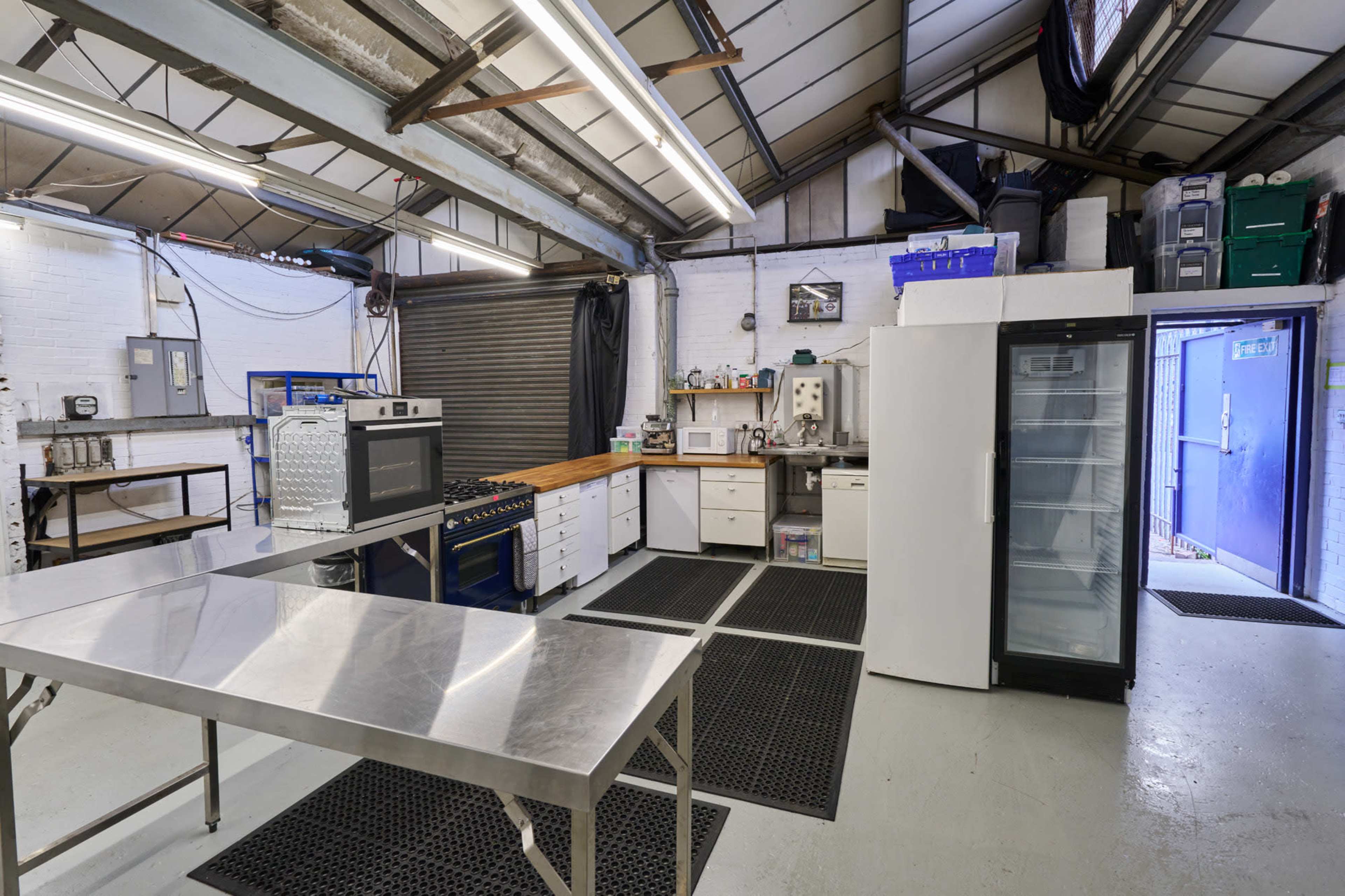 The image shows a commercial kitchen with stainless steel tables, an oven, a refrigerator, and various kitchen appliances against a white brick wall.
