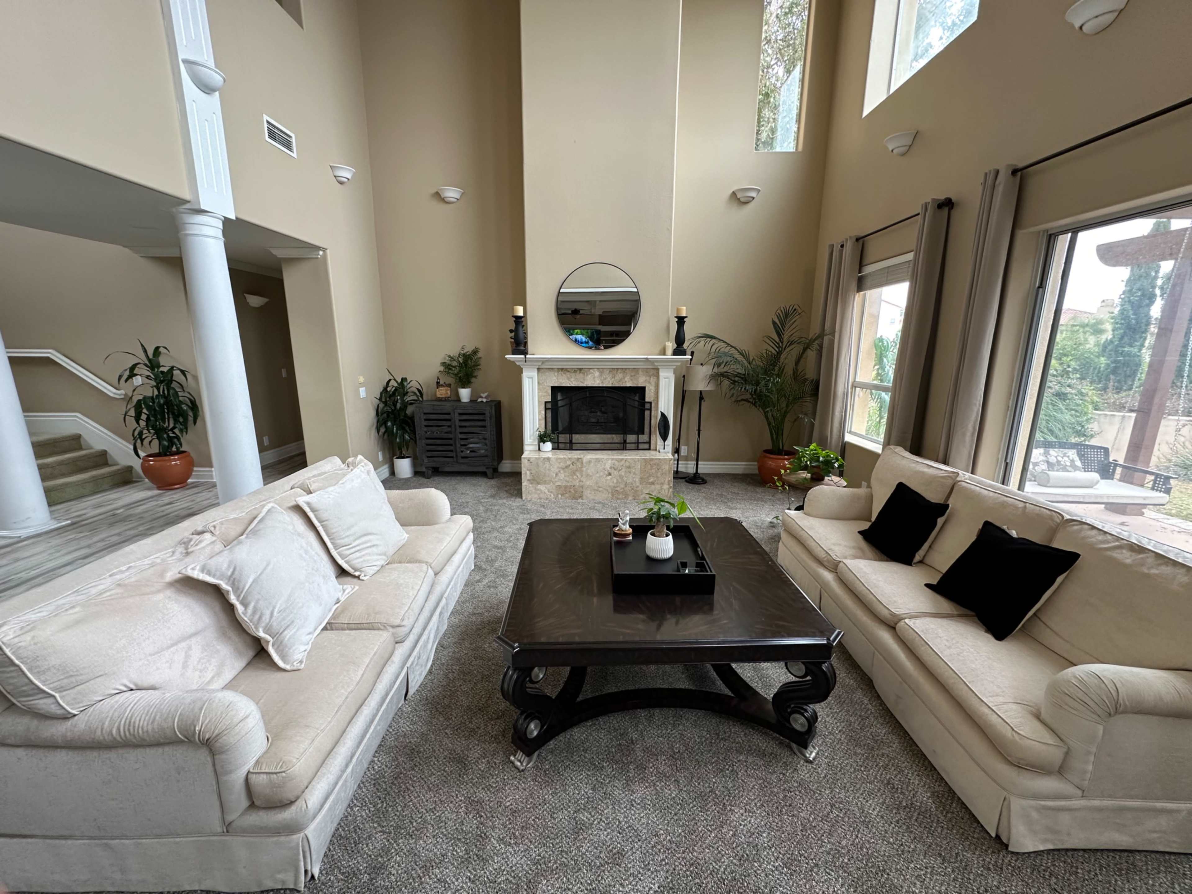 The living room features two beige sofas facing a dark wooden coffee table, with a marble fireplace and large windows allowing natural light in.