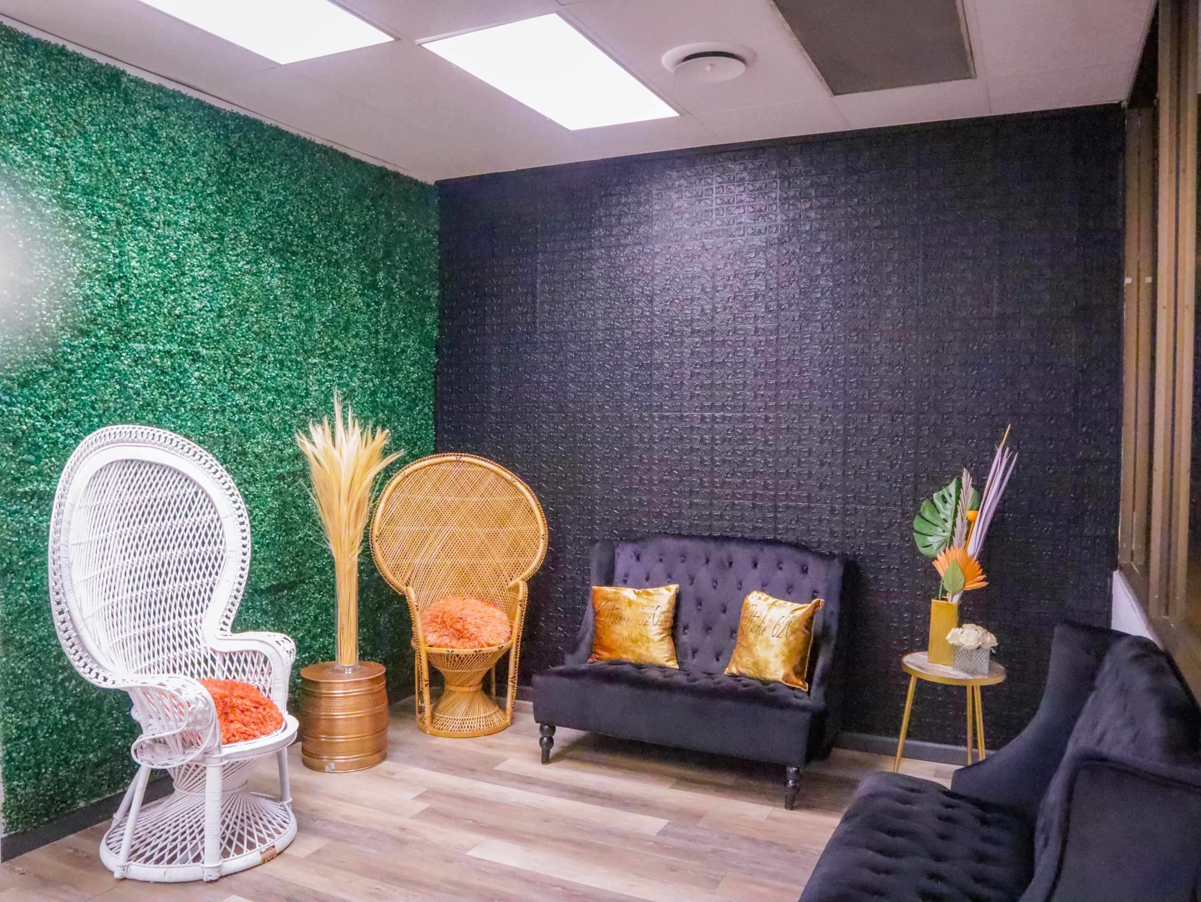 The room features a combination of green artificial grass on one wall and a black textured wall, furnished with two ornate chairs, a sleek black sofa, and decorative plants.