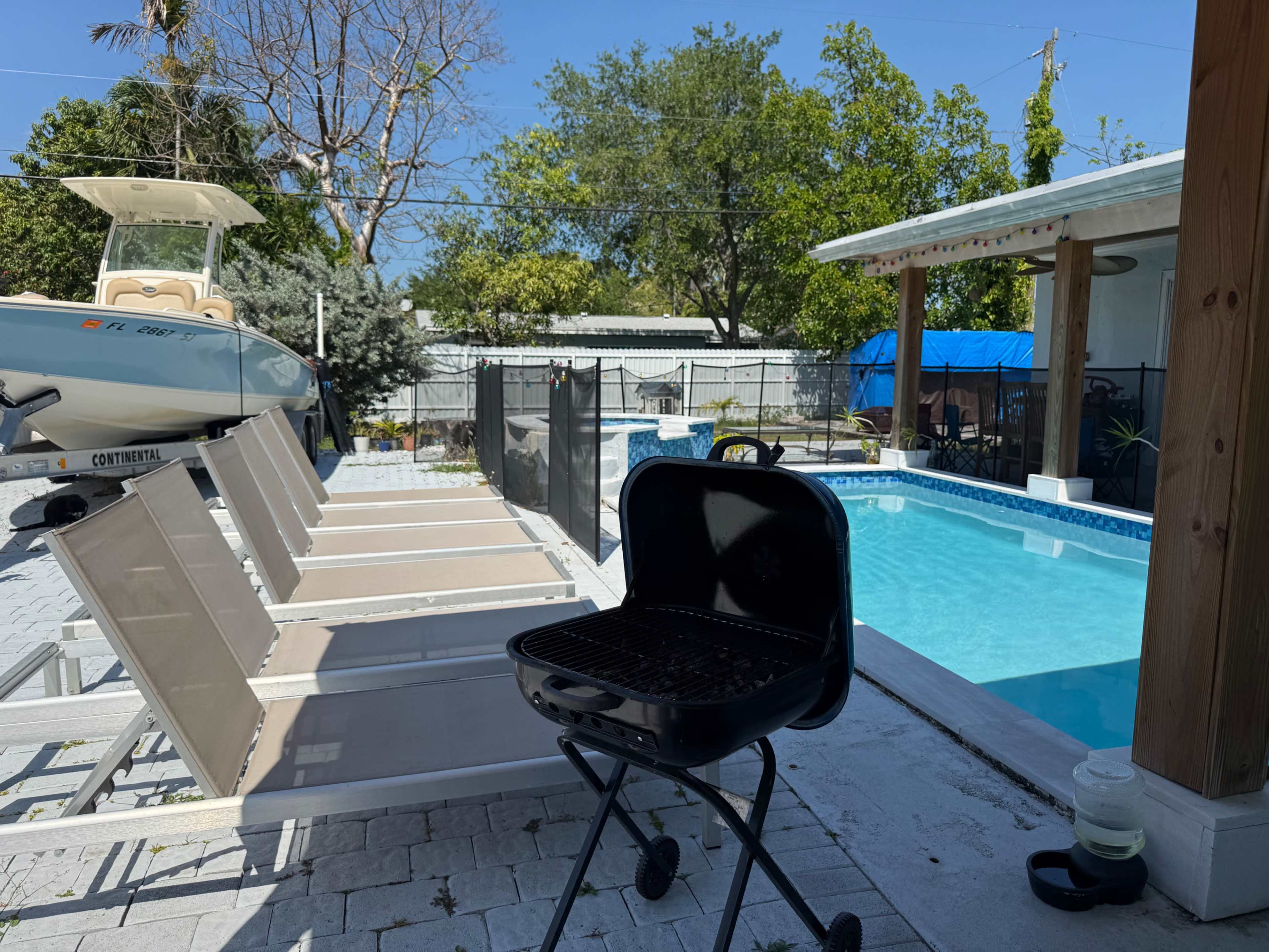 The image shows a backyard with a swimming pool, lounge chairs lined up beside it, and a grill nearby, under a clear blue sky.