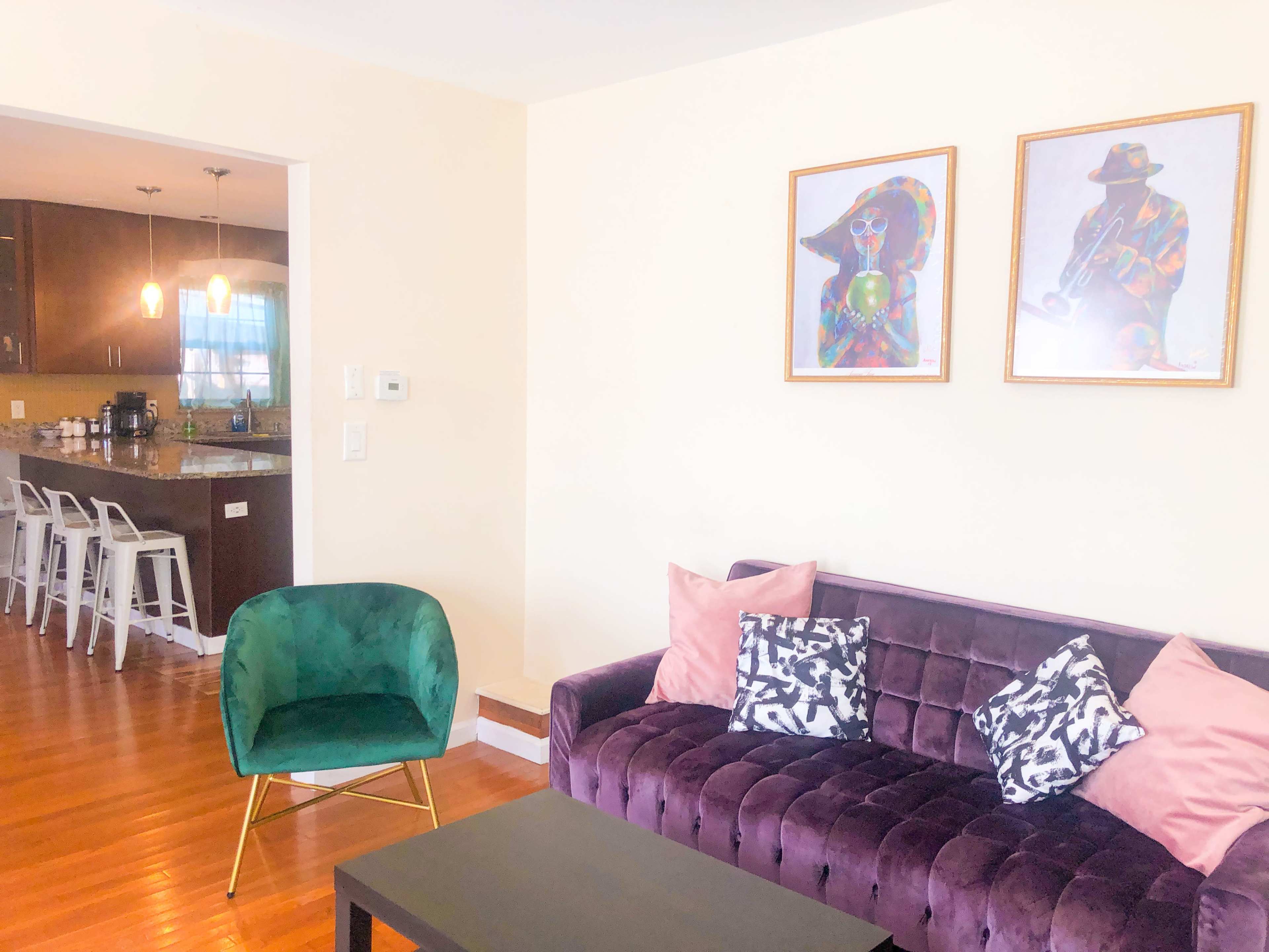 The image shows a living room with a purple sofa adorned with decorative pillows, a green chair, a coffee table, and a view of a kitchen in the background with bar stools and pendant lights.