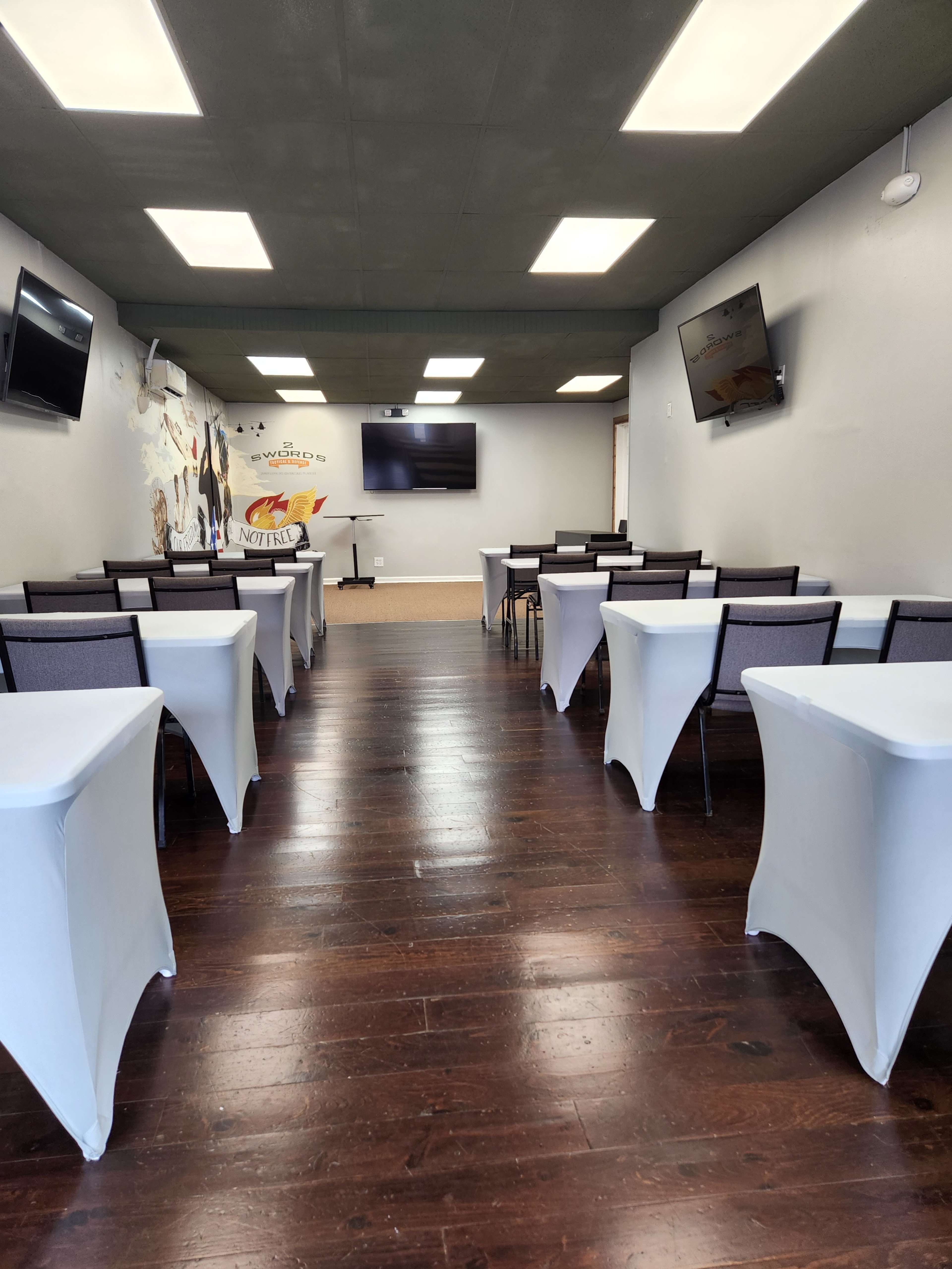 A classroom or meeting space with rows of tables covered in white tablecloths, equipped with wall-mounted televisions and a mural on one wall.