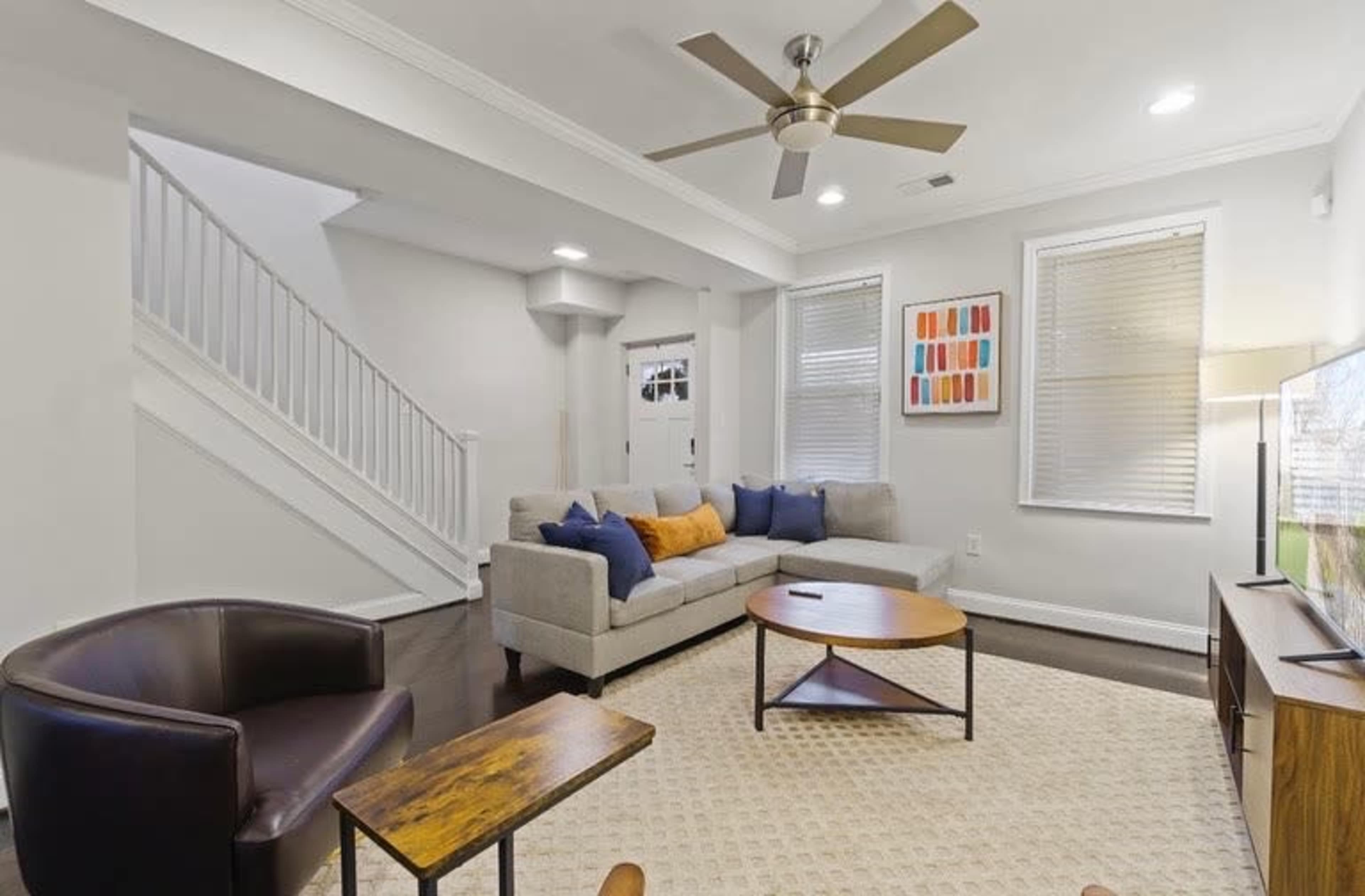 The living room features a gray sofa with blue and orange pillows, a round wooden coffee table, and a television, with a staircase visible in the background.