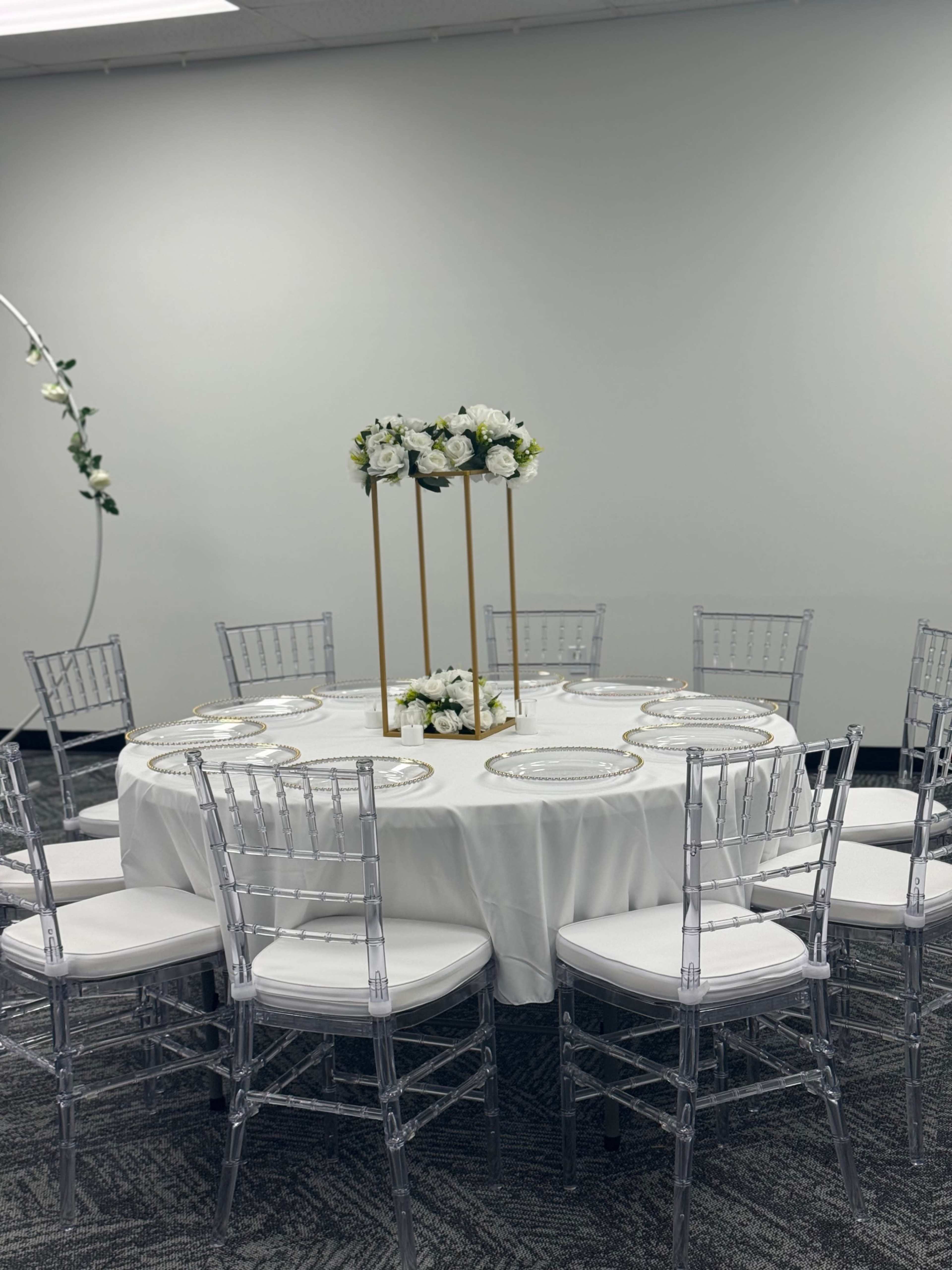 A round table is set with white linens and elegant decor, including floral arrangements atop tall stands, surrounded by clear chiavari chairs.
