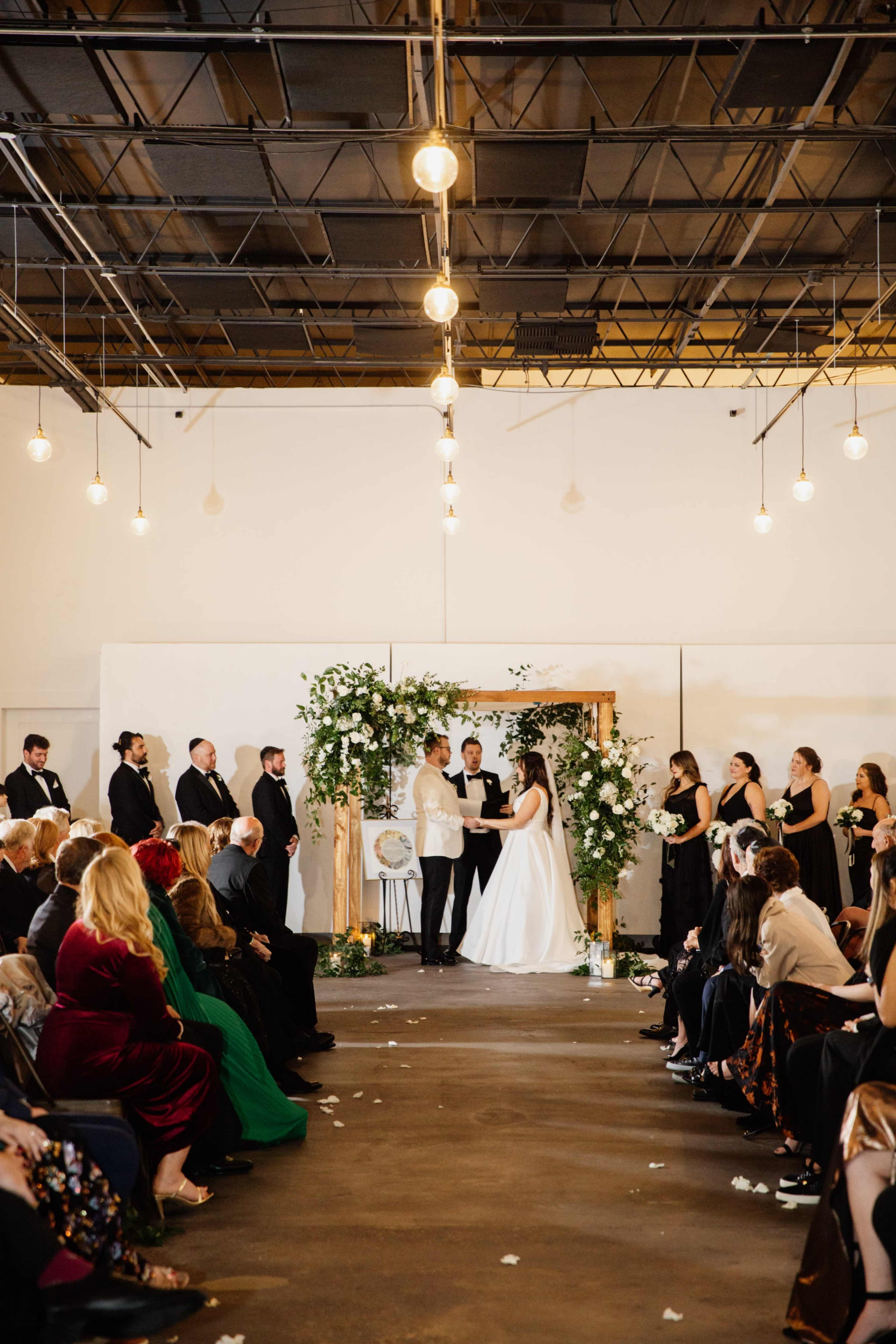 A couple exchanges vows under a floral arch during a wedding ceremony, while guests seated on either side observe the event.