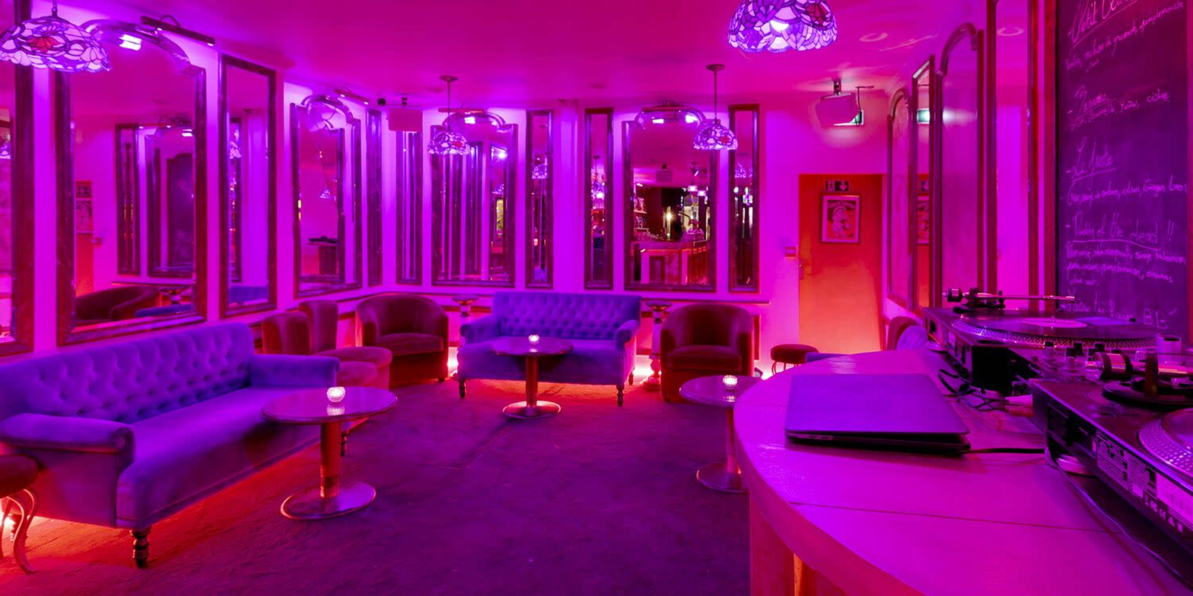 A lounge area with purple lighting features plush seating, mirrored walls, and a DJ setup.