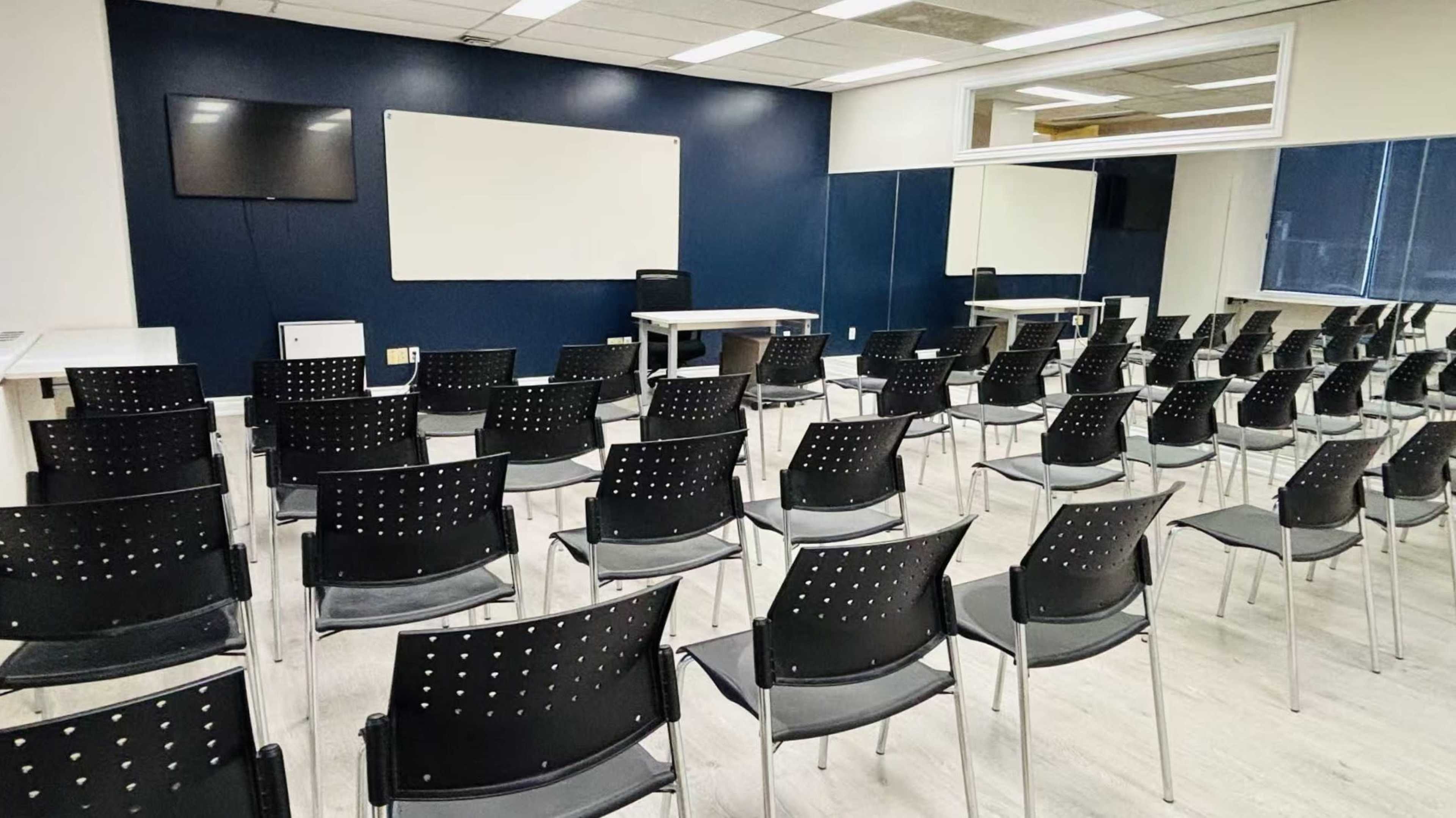 The image shows a classroom with rows of black chairs facing a whiteboard and a wall-mounted television in a well-lit environment.