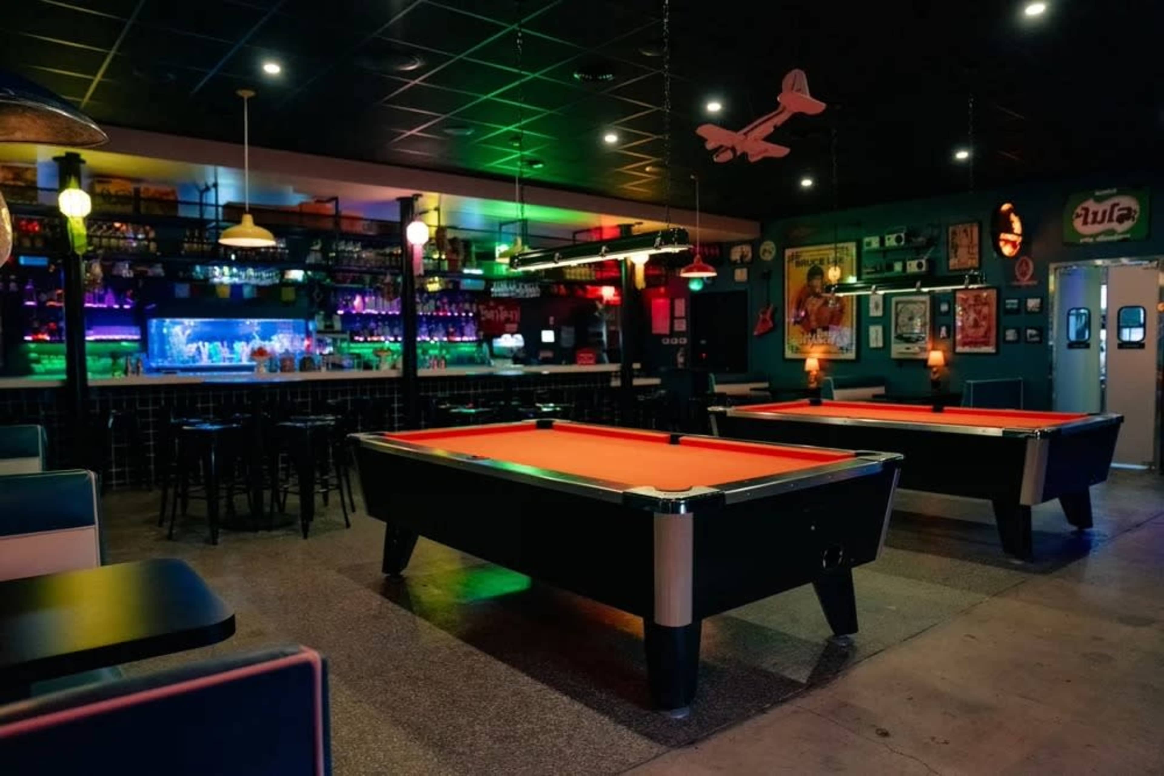 The image shows a dimly lit bar with several pool tables, a colorful backlit bar, and various decor on the walls.