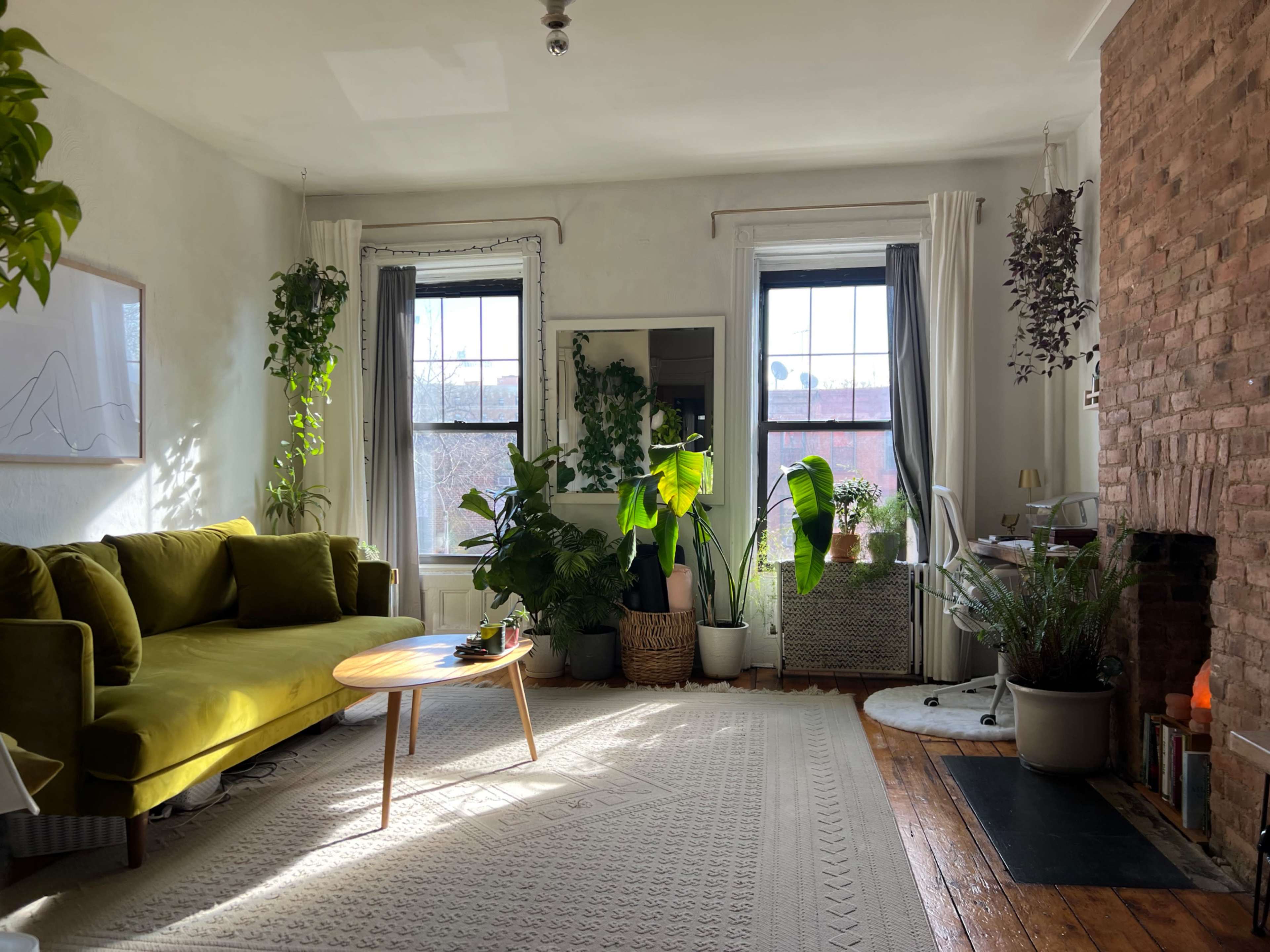 A cozy living room features a green sofa, large windows with plants, and a brick fireplace.