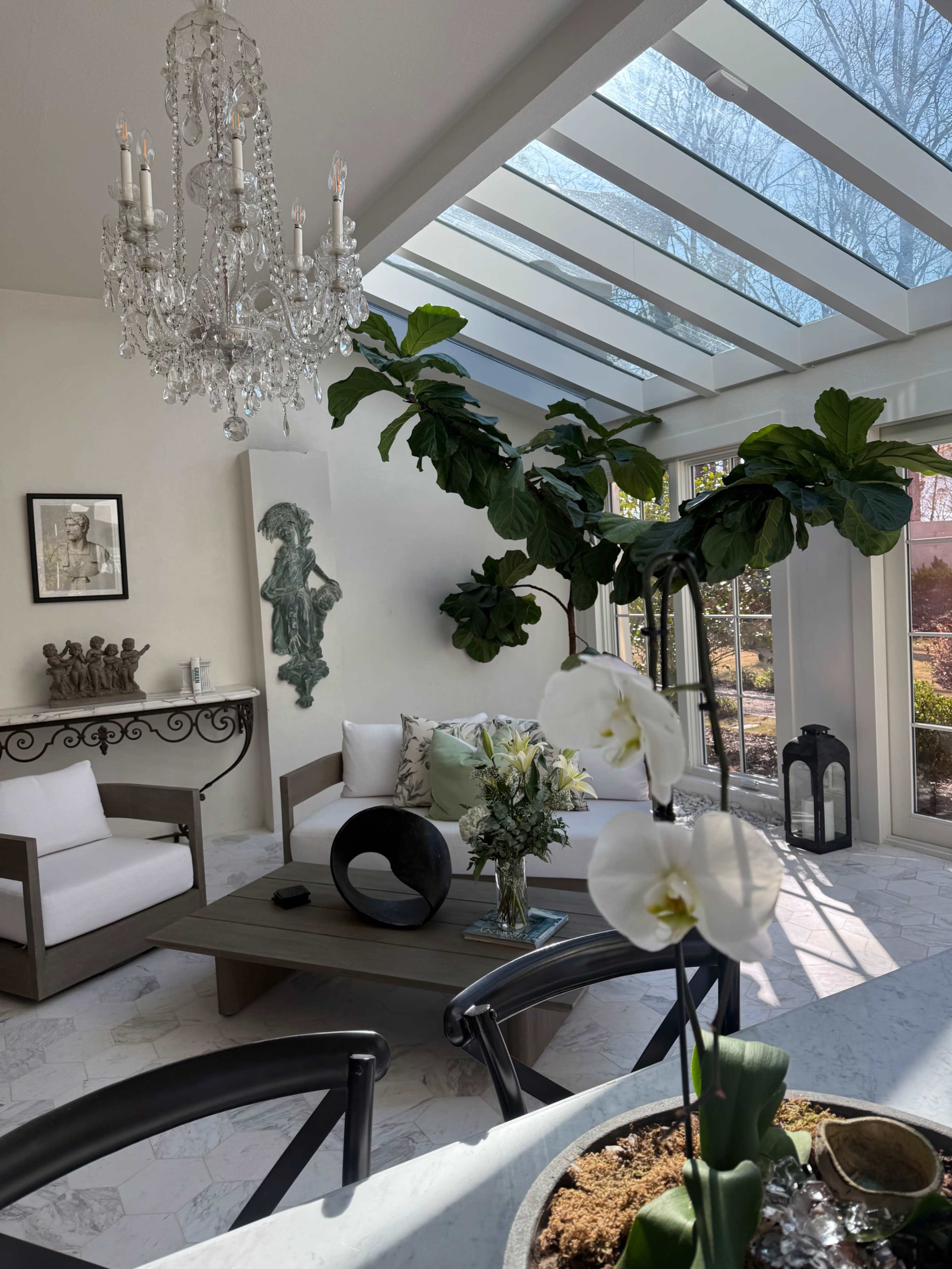 The image shows a bright, airy sunroom with a chandelier, large windows, indoor plants, and contemporary furniture.