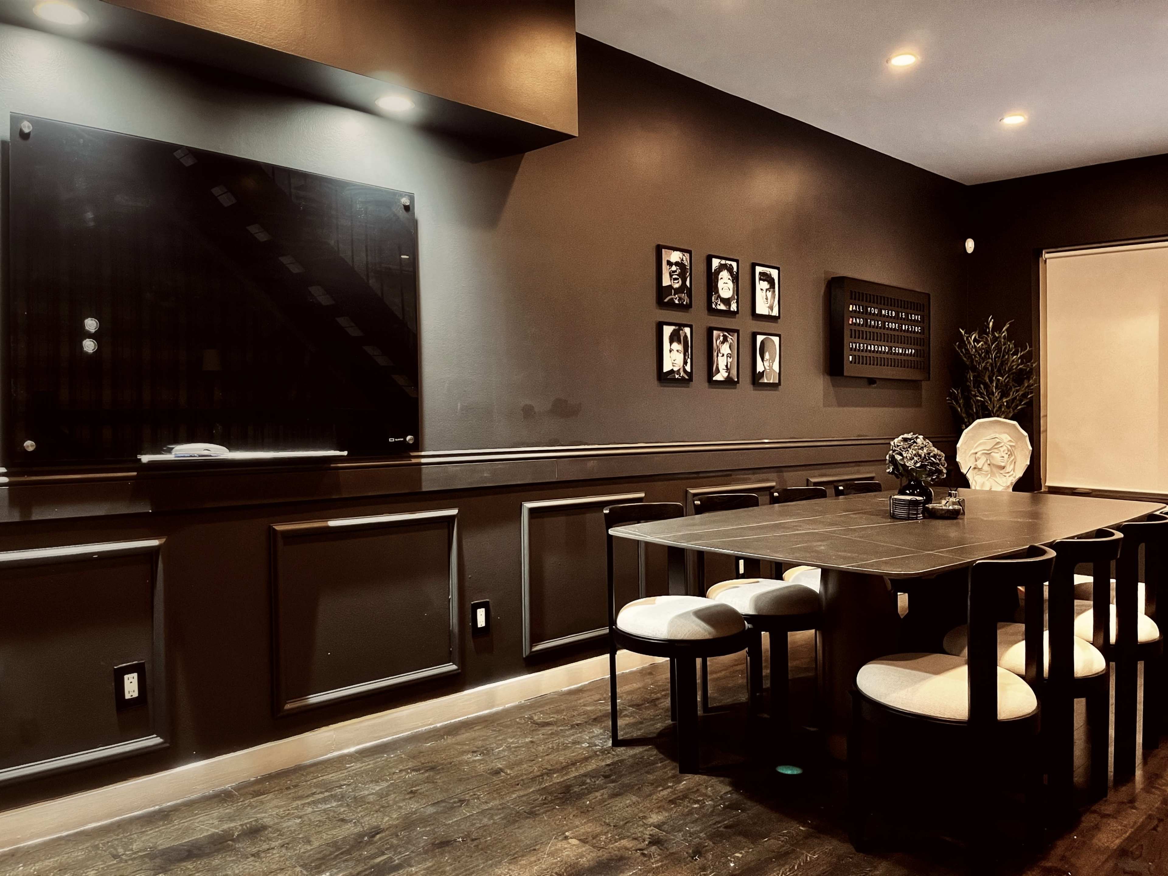 A dark-colored dining room features a long table surrounded by six chairs, with a television mounted on the wall and framed photographs displayed nearby.