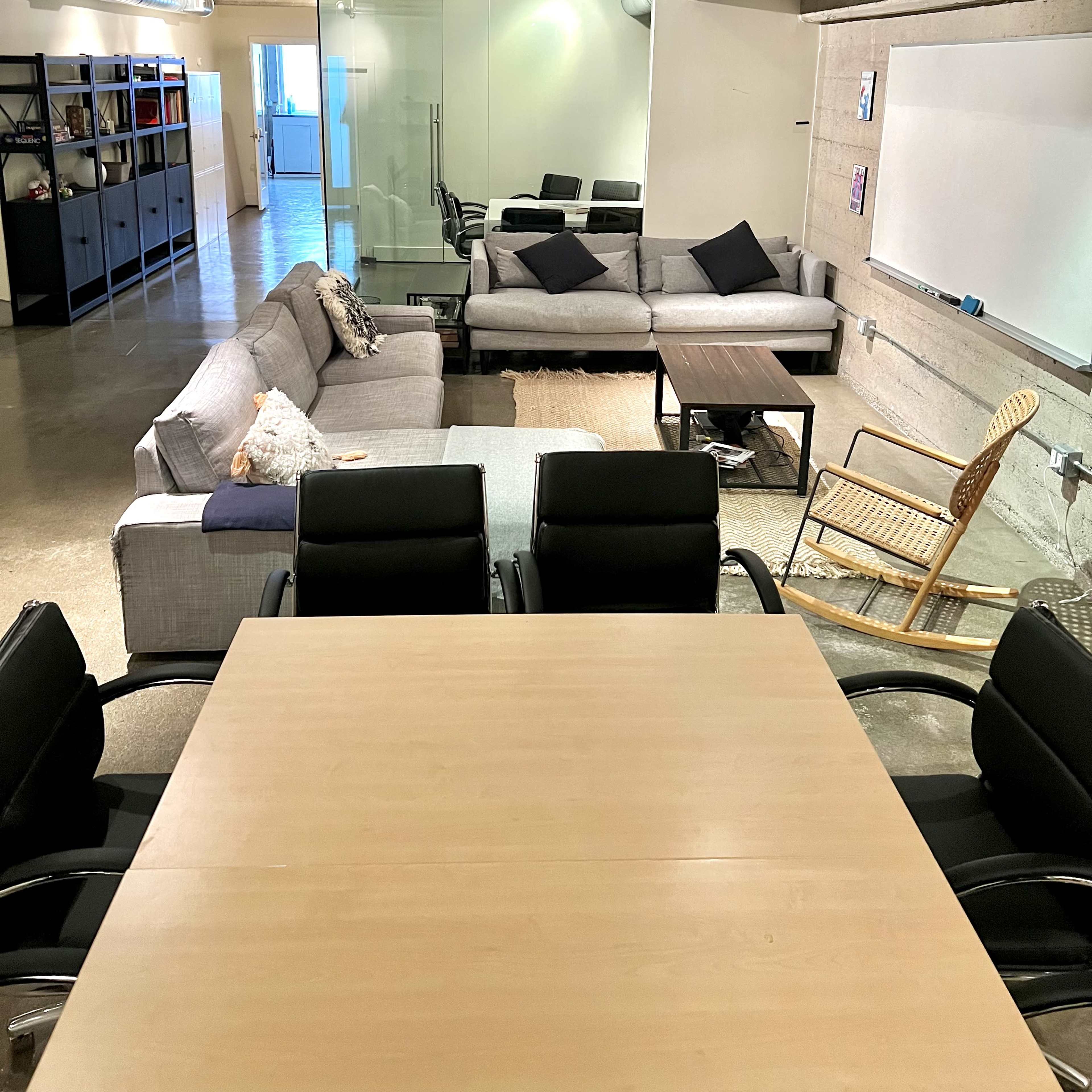 A spacious modern lounge area with multiple seating arrangements, including sofas, a coffee table, and a rocking chair, alongside a conference table with black office chairs.