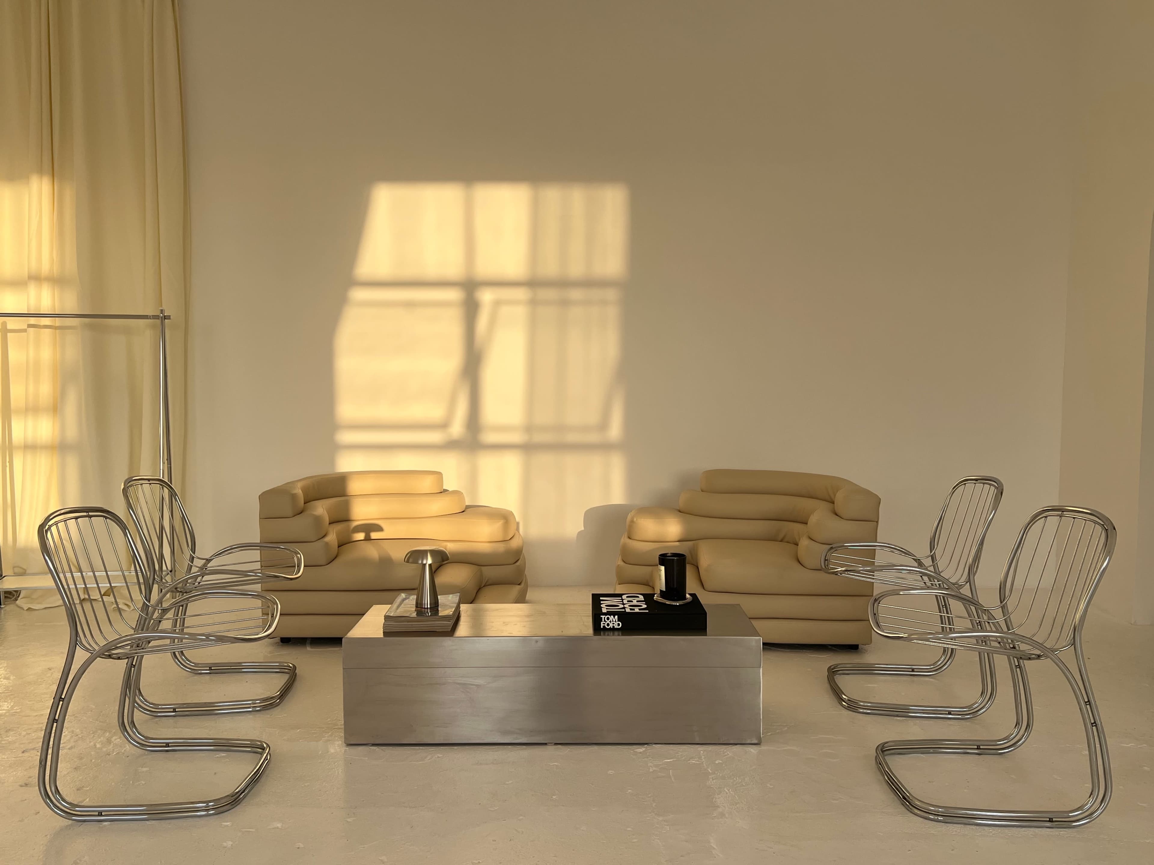 A minimalist seating area with two pale sofas facing each other, surrounded by six metallic chairs, and a rectangular coffee table in the center.